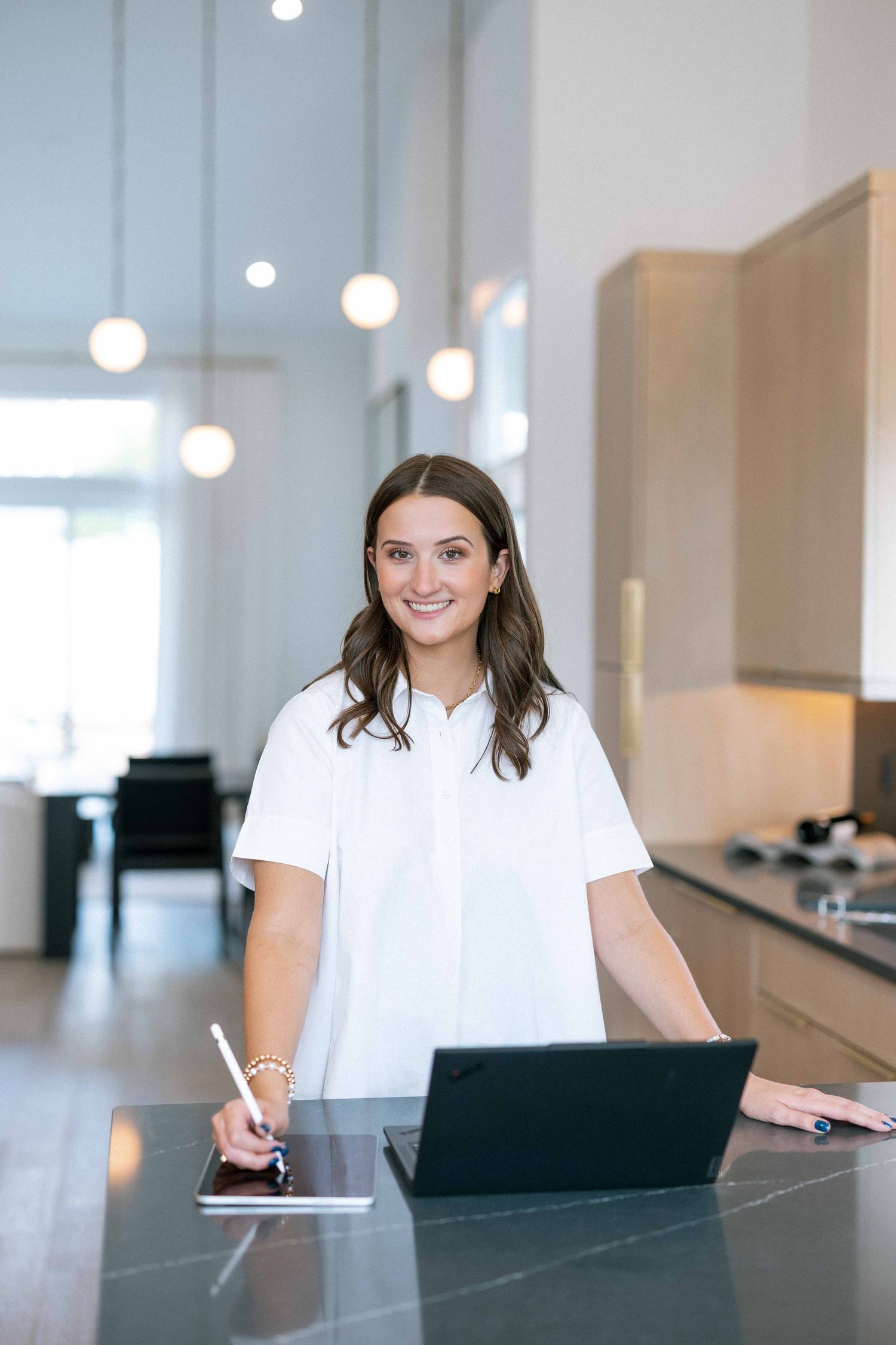 Smiling brunette woman in a white blouse working at a modern kitchen counter with a laptop and tablet.