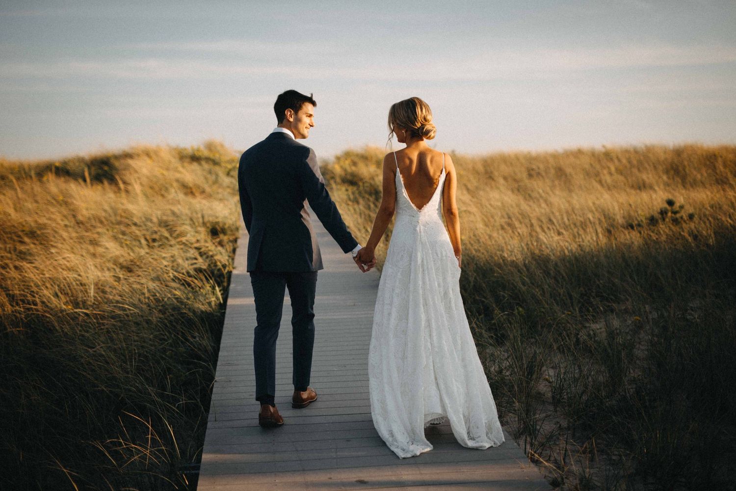 Bride and groom walk hand-in-hand on a wooden boardwalk through golden beach grass at sunset.