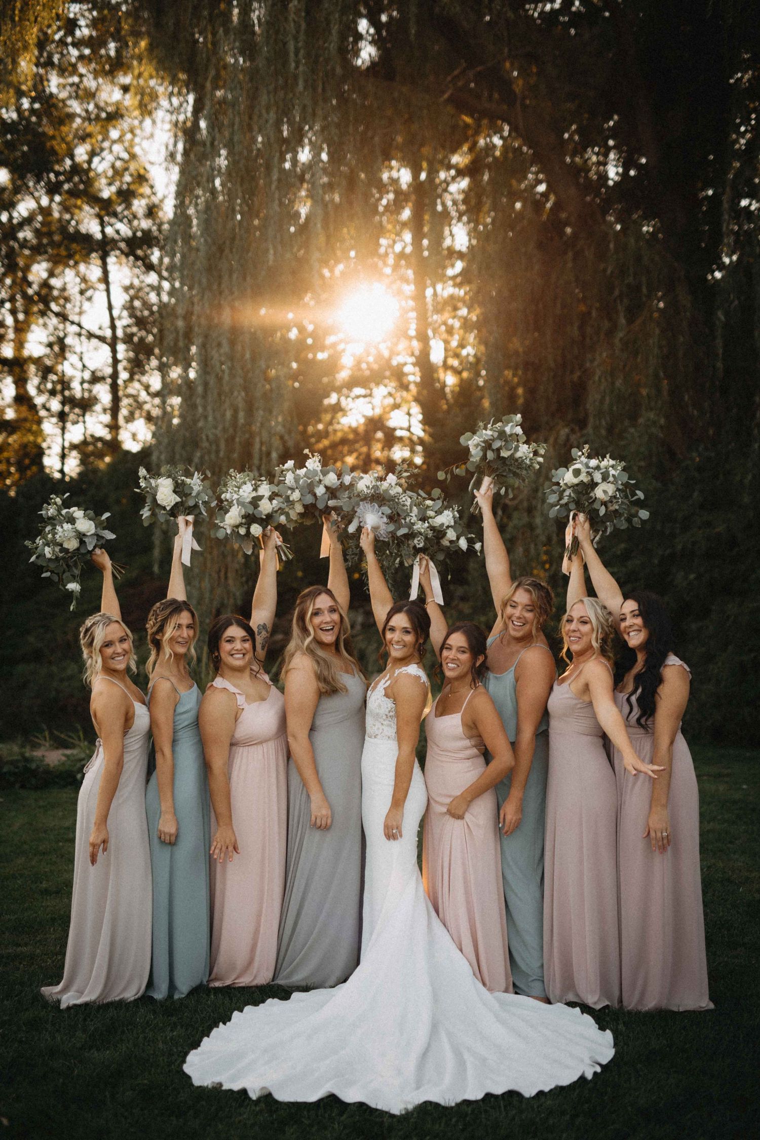 Bridesmaids in mismatched blush and sage gowns raise bouquets with bride in golden sunset light beneath tall trees.