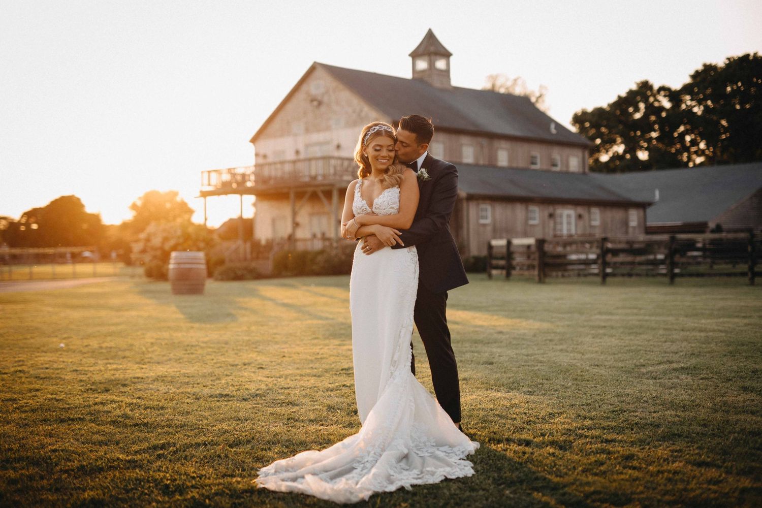 Groom embracing bride in lace gown during golden hour portrait at rustic barn wedding venue.