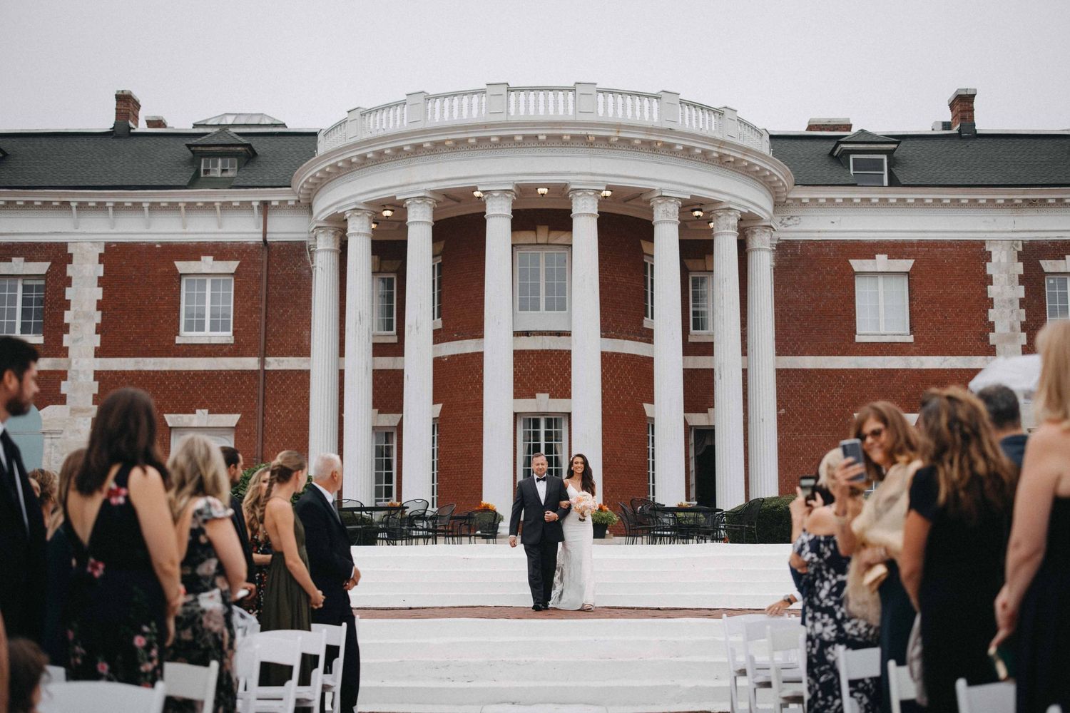Groom walks bride down the aisle at an outdoor ceremony before a grand brick estate.