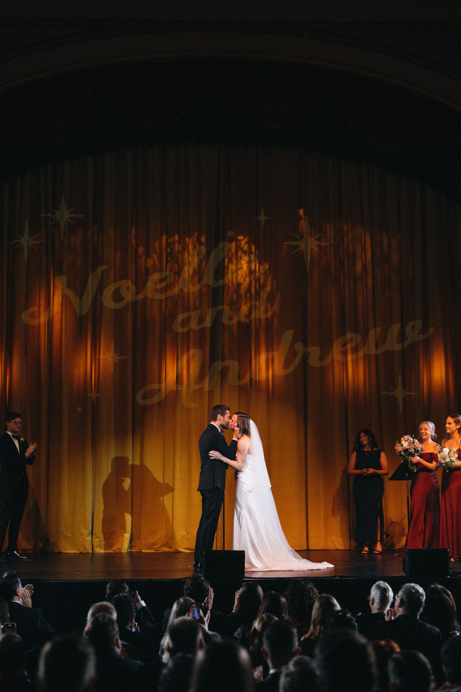 Couple shares first kiss on stage with names projected on golden curtain at wedding ceremony.