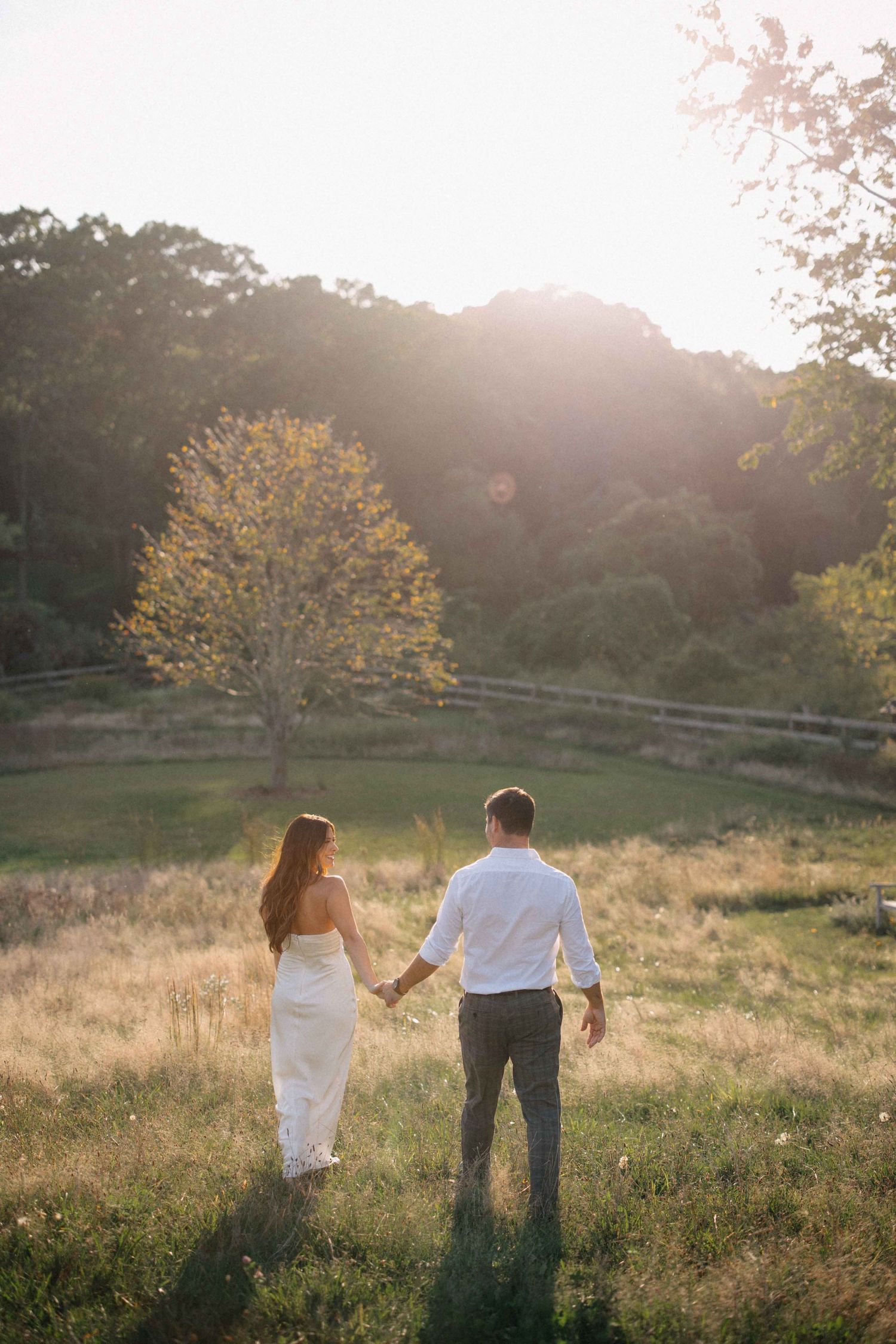 A couple holds hands walking through a sunlit golden meadow with rolling hills and trees behind them.