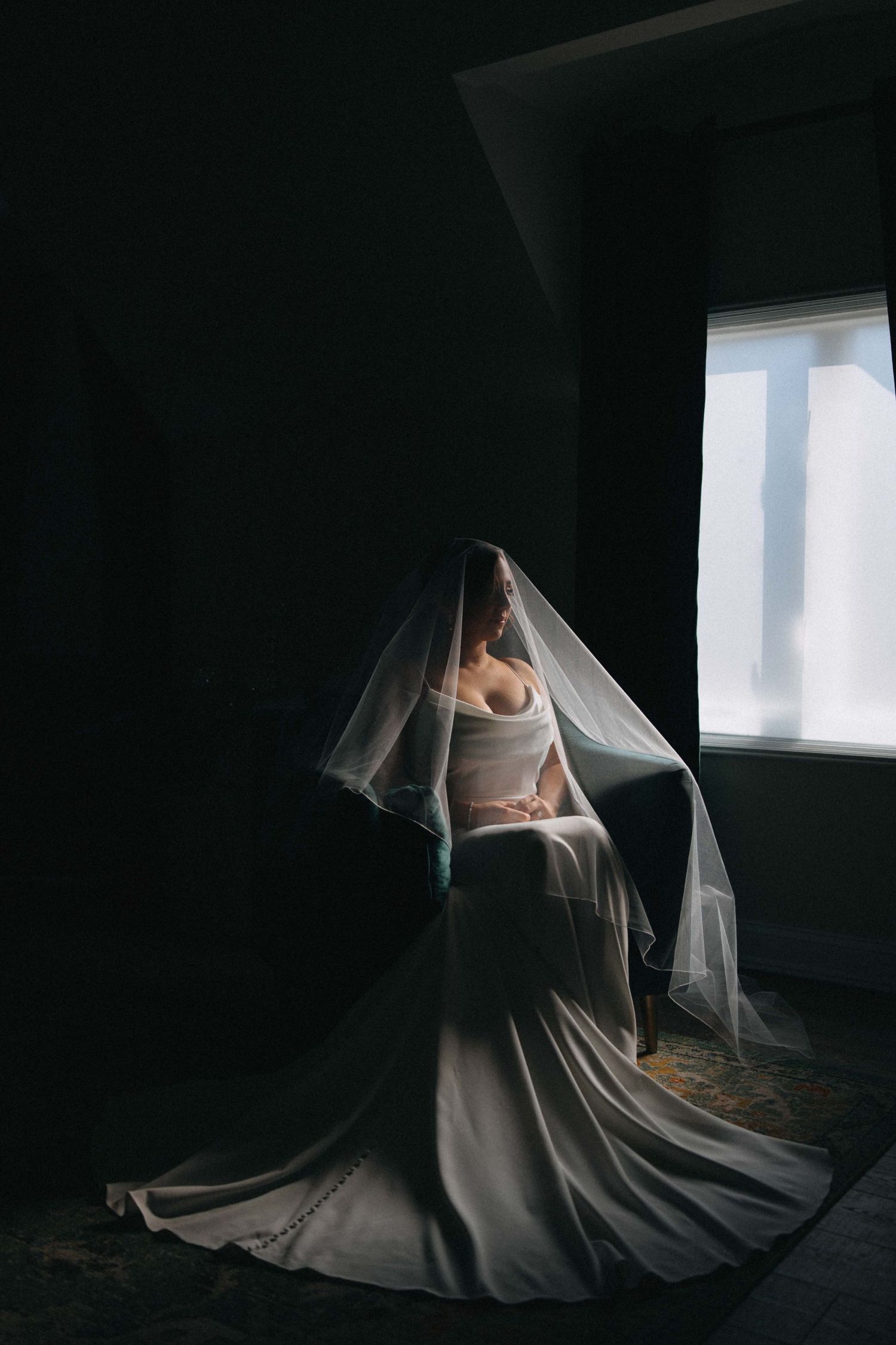 Bride in wedding gown covered by veil seated by bright window in dark room.