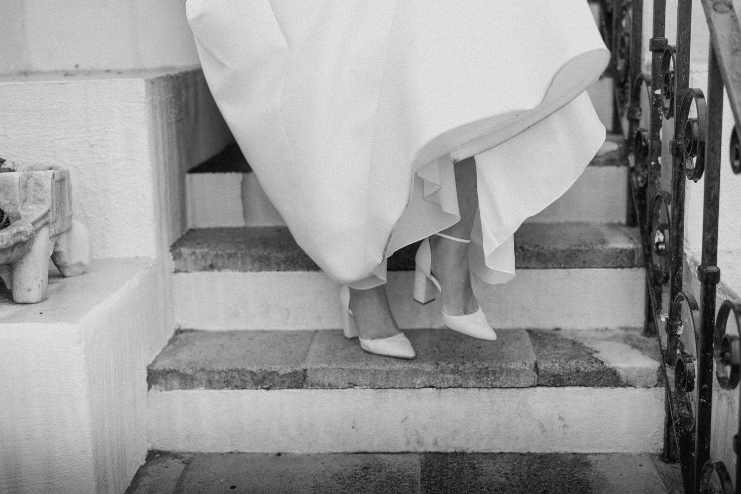 Black and white photo of bride in heels ascending stone steps with iron railing.