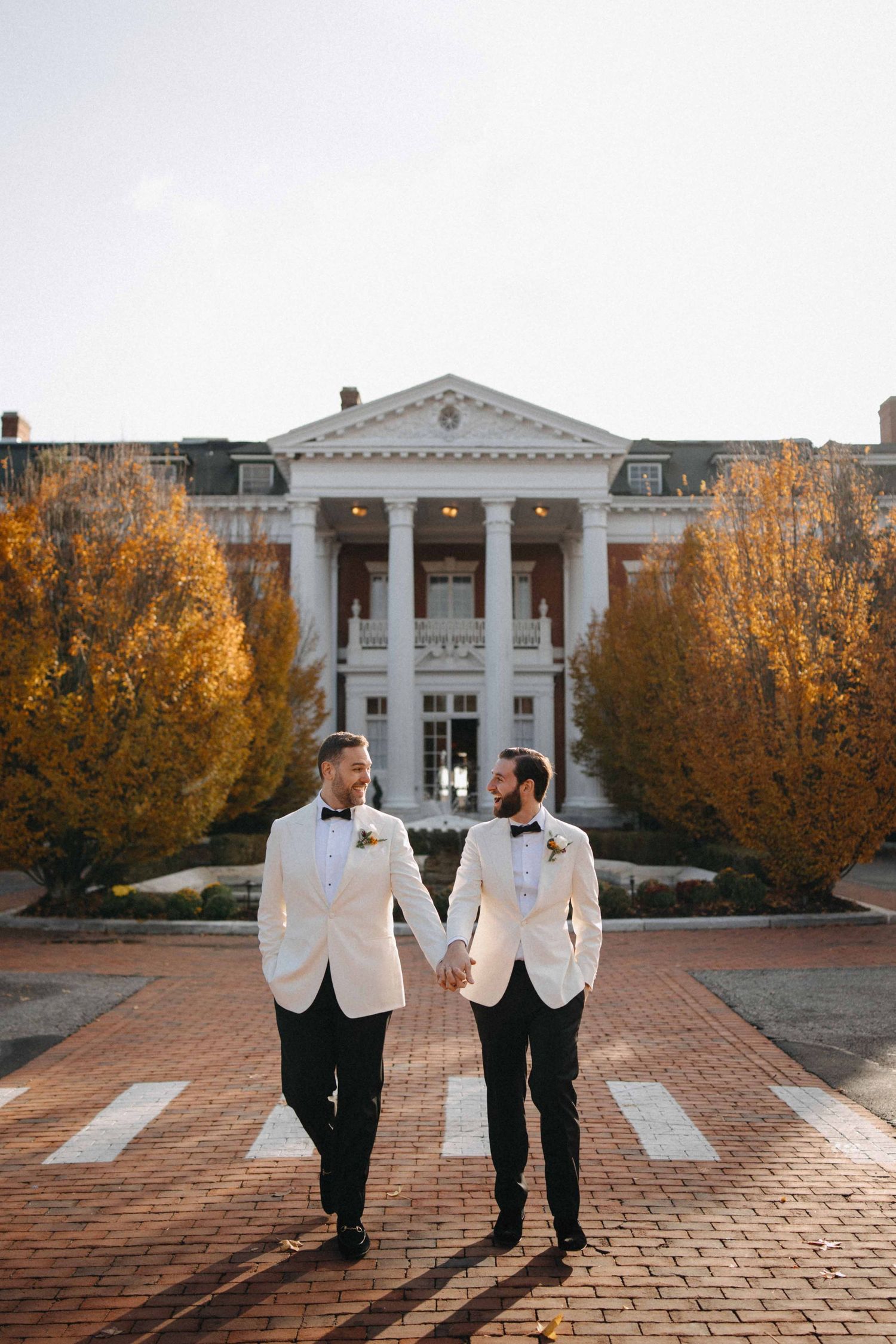 Two grooms in white tuxedo jackets hold hands on a brick path before a grand white columned building in autumn.