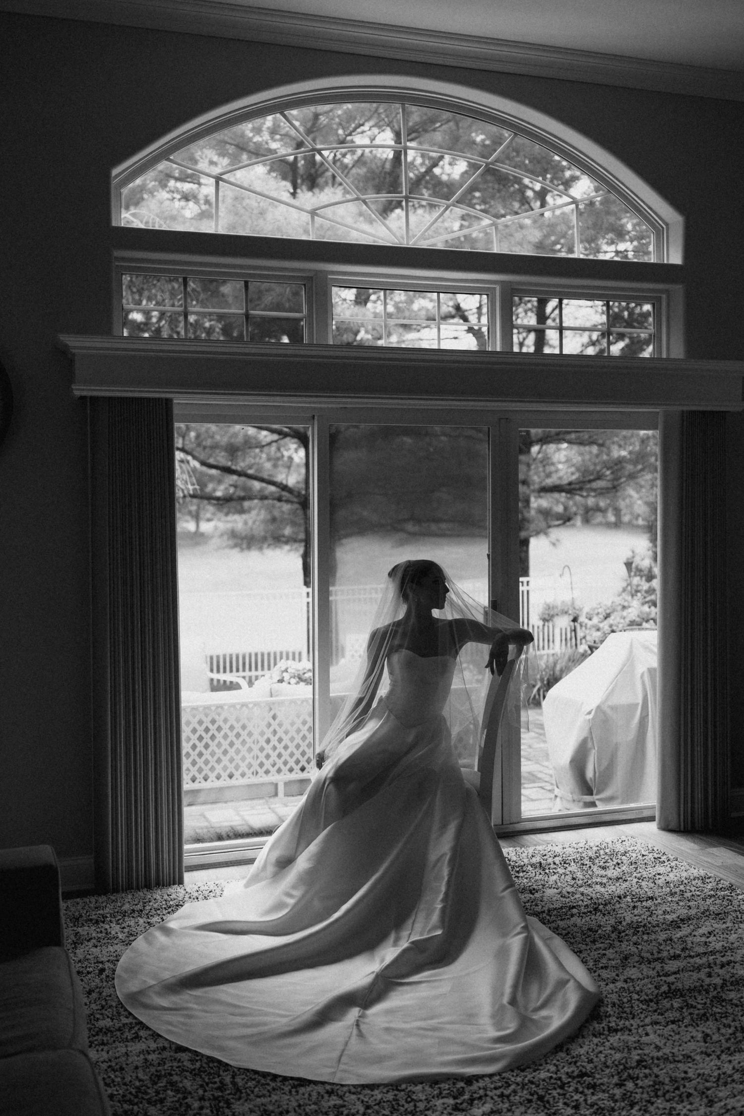 Black and white photo of a bride in a flowing gown and veil standing before large French doors with arched window.