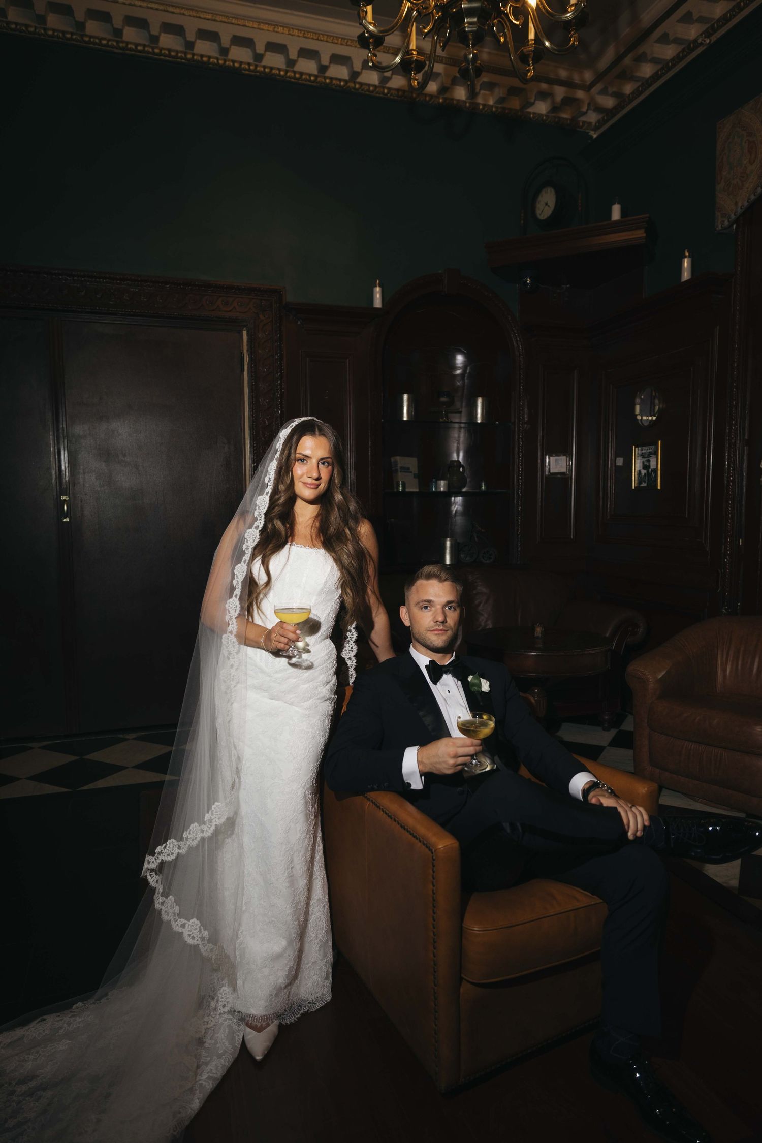 Bride in white lace gown and veil stands beside groom in tuxedo holding cocktails in elegant dark interior.