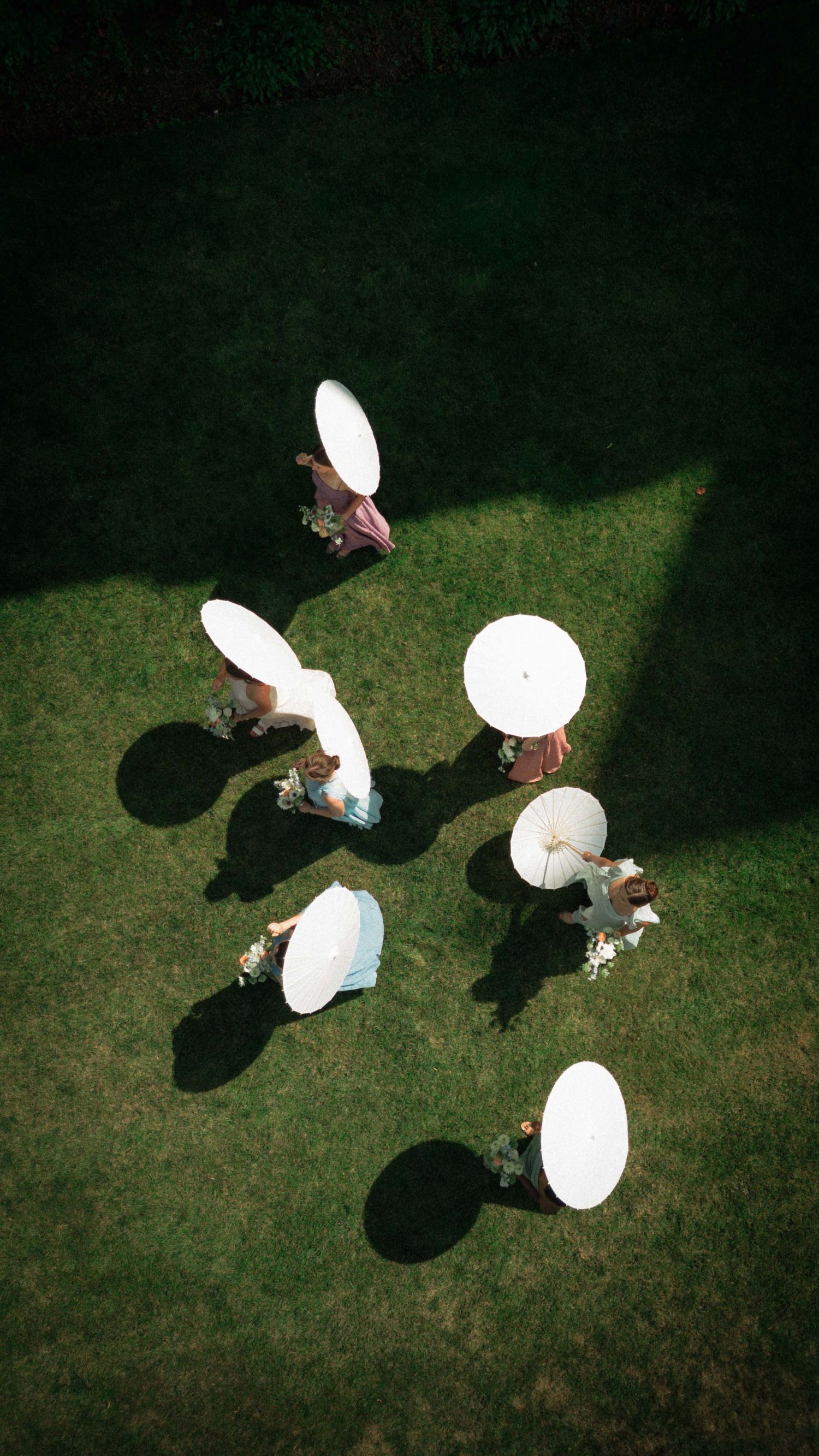 Aerial view of people holding white parasol umbrellas on green grass, casting shadows in natural sunlight.