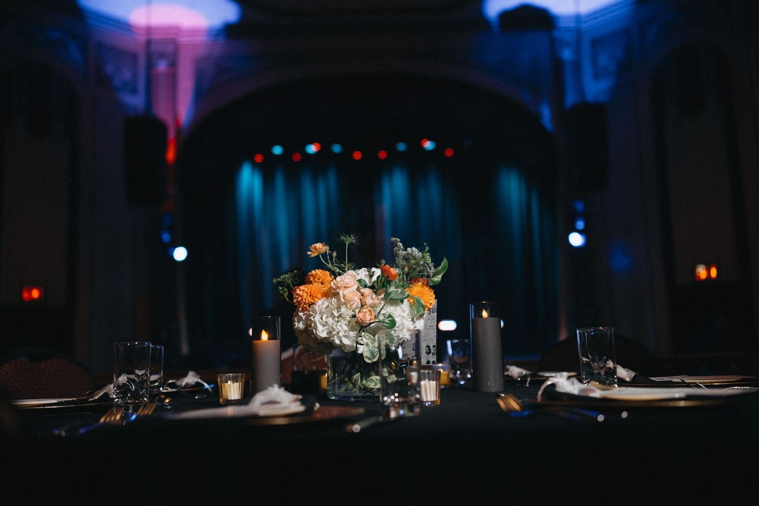 Orange and white floral centerpiece with candles on a dark reception table with a stage backdrop.