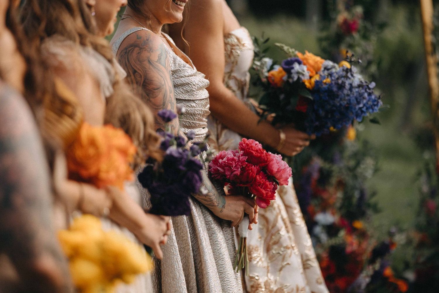 Bridesmaids holding colorful wedding bouquets of red, purple, blue, and orange flowers at an outdoor ceremony.
