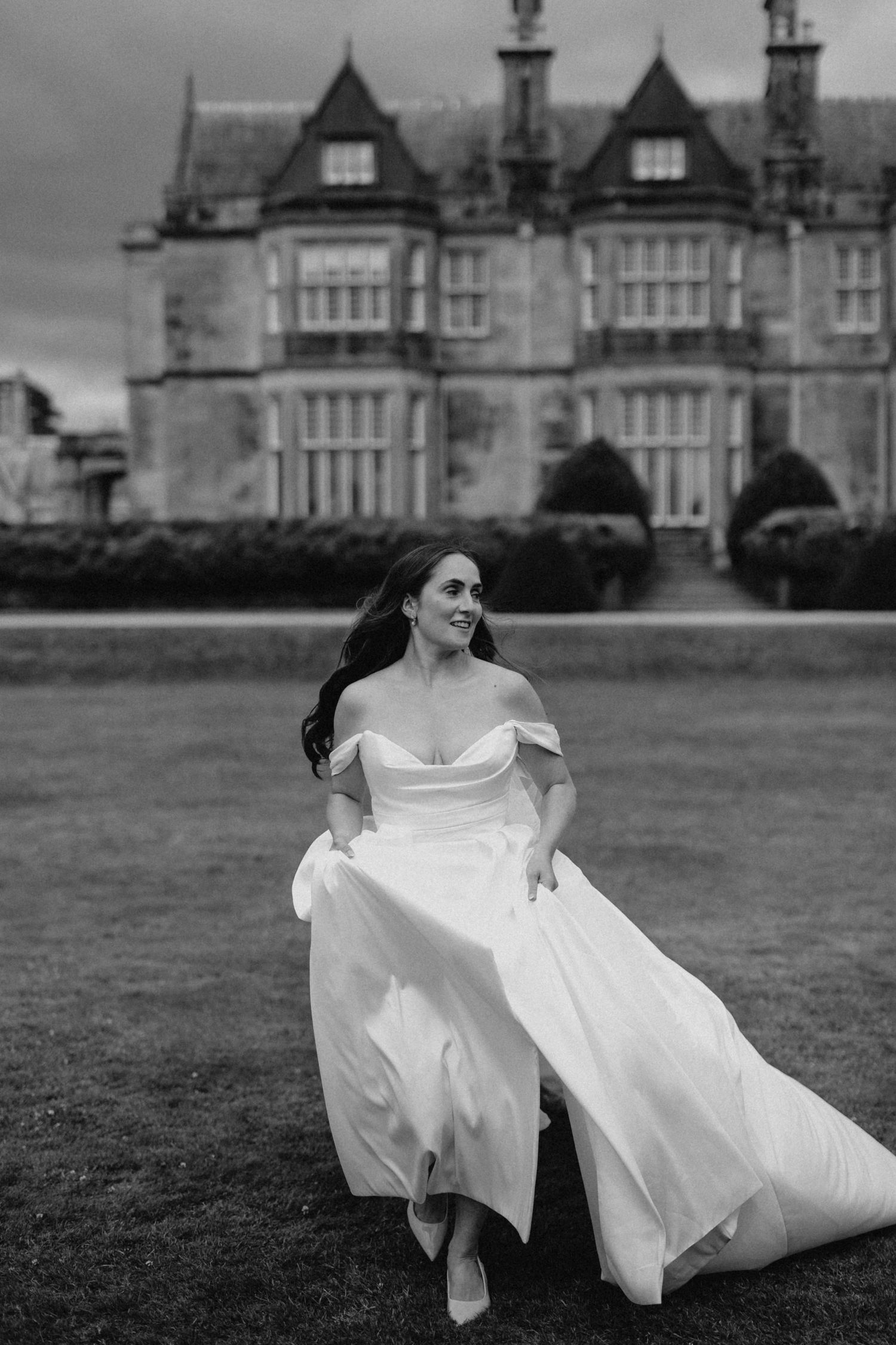 Bride in off-shoulder white gown runs across lawn before grand estate in black and white wedding photo.