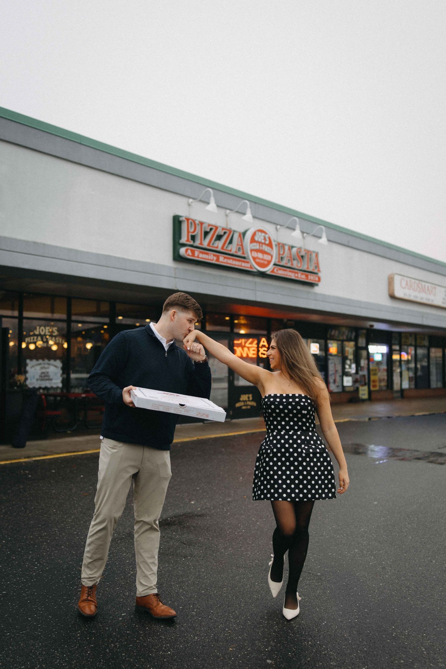 Couple outside Pizza Diana restaurant, woman in polka dot dress feeding man holding pizza box.