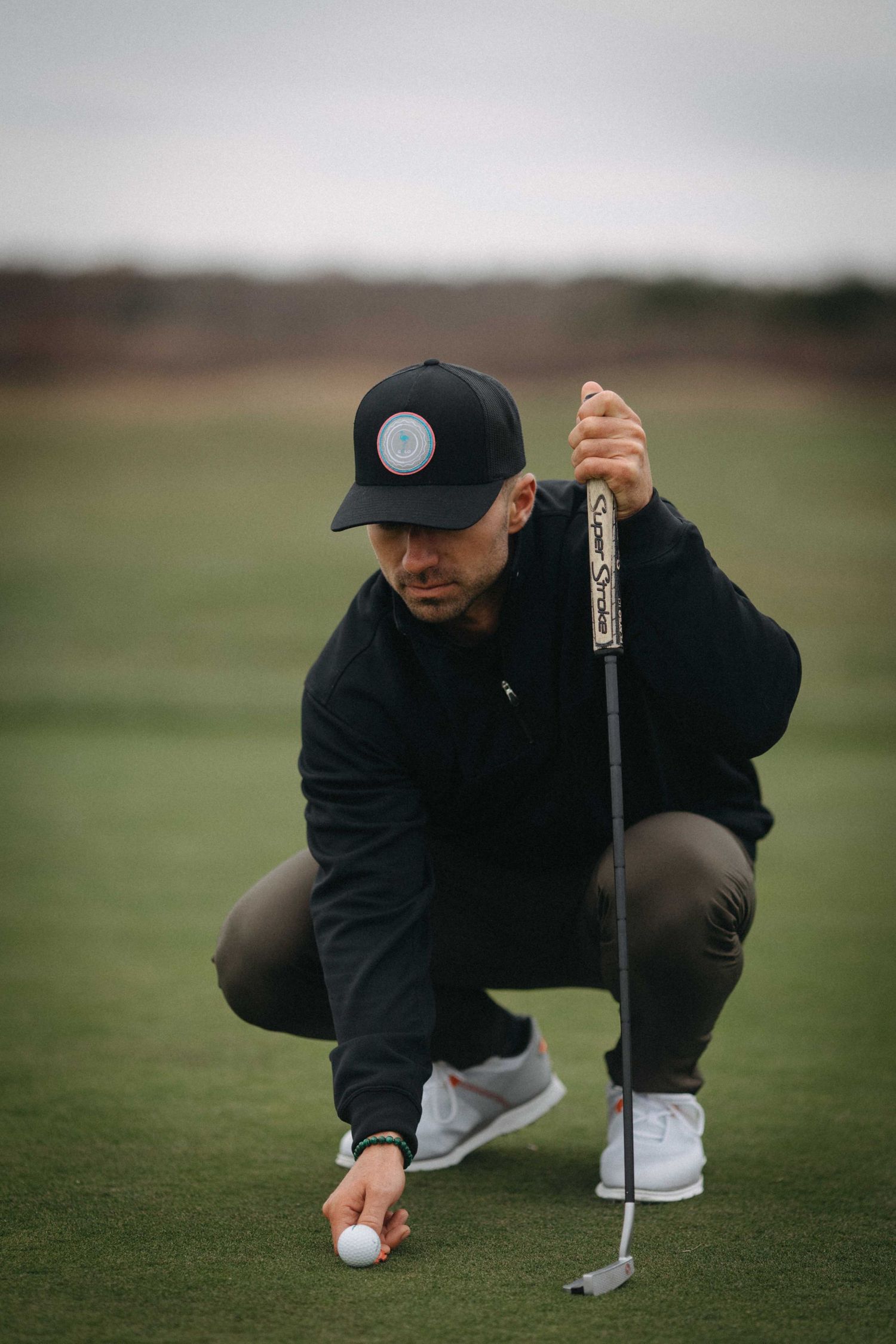 Male golfer in black outfit crouching to place golf ball on green with putter.