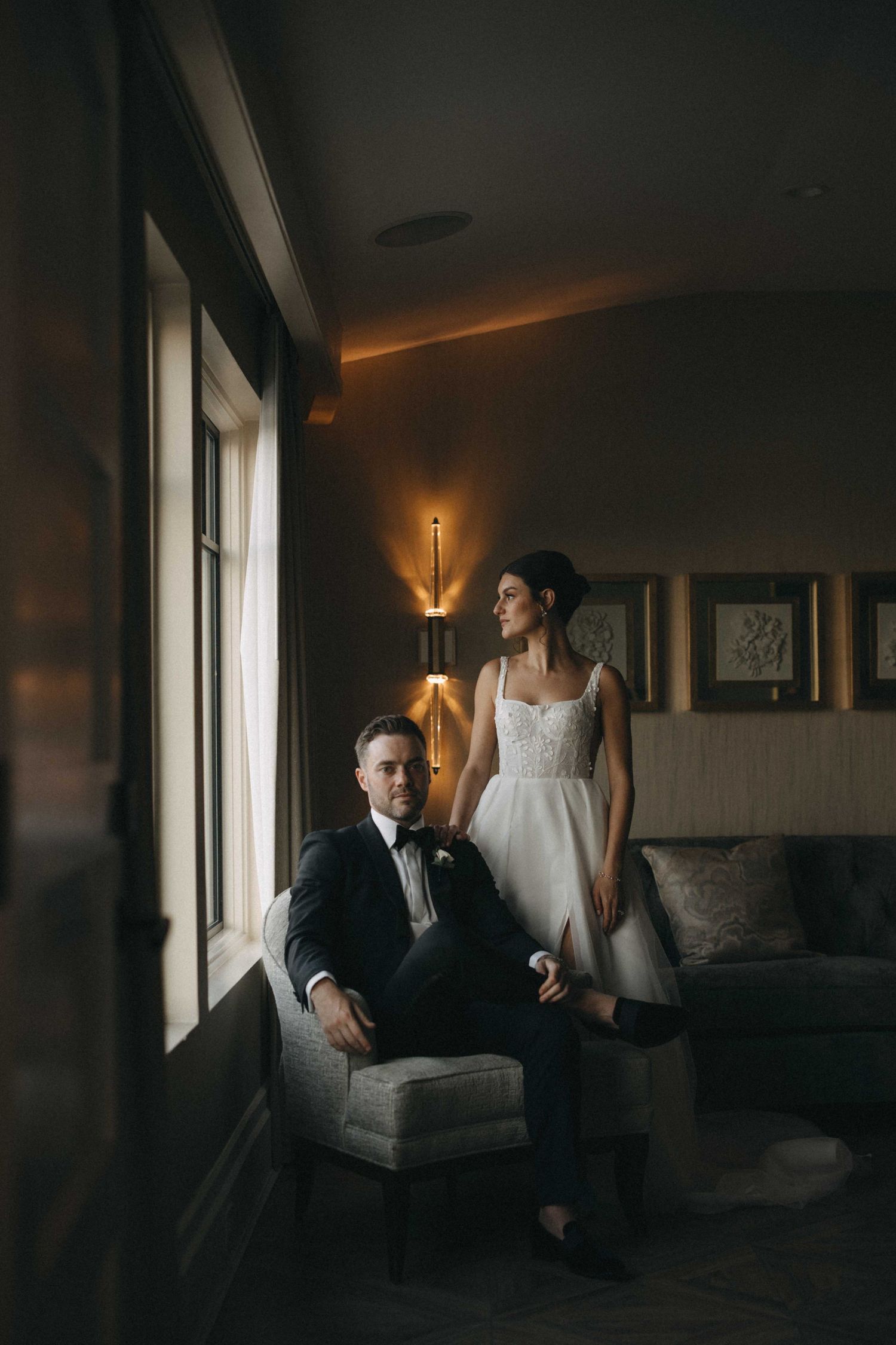 Bride and groom posing elegantly by window in dimly lit luxury hotel room.
