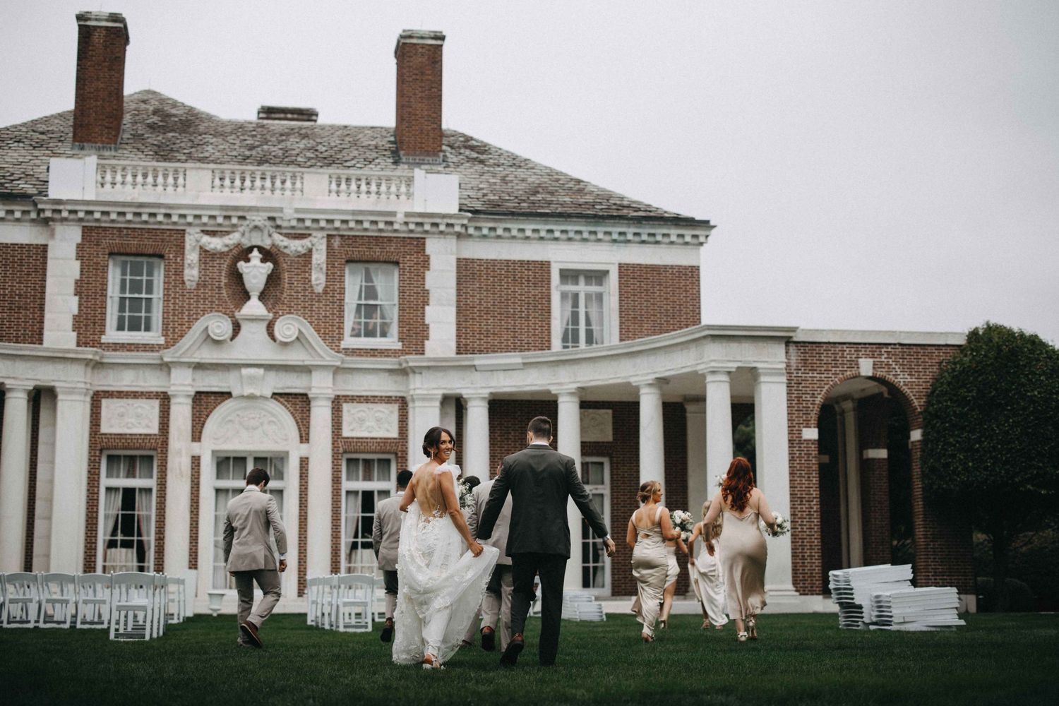 Bride, groom, and wedding party walk toward grand brick colonial mansion across manicured lawn with white chairs.