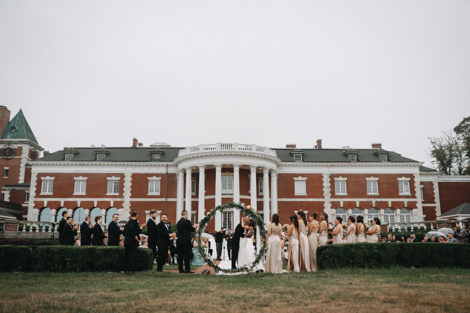 Wedding party poses in front of a grand brick colonial estate with white columns.