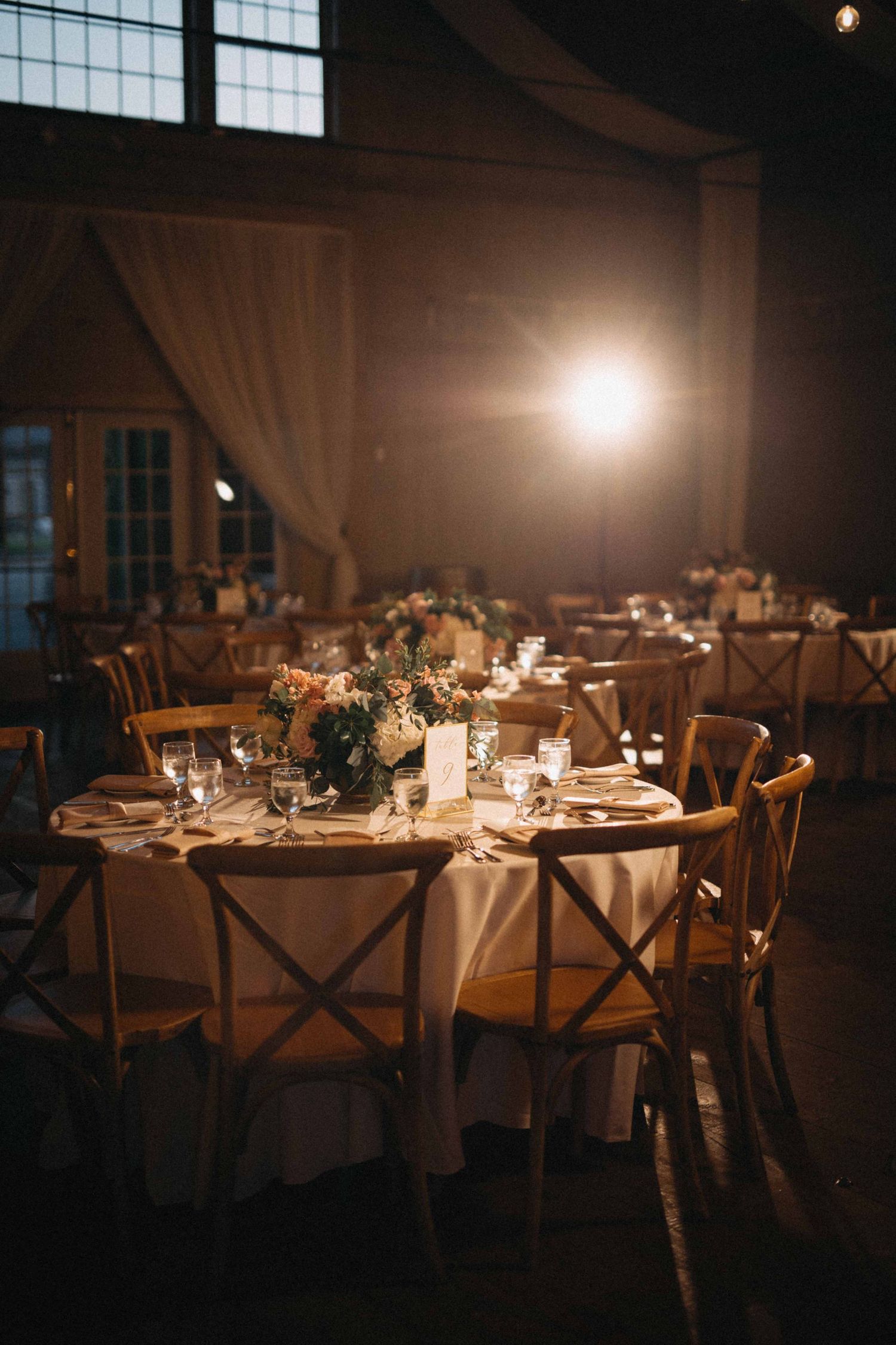 Warmly lit barn wedding reception with wooden cross-back chairs and floral centerpieces on round tables.