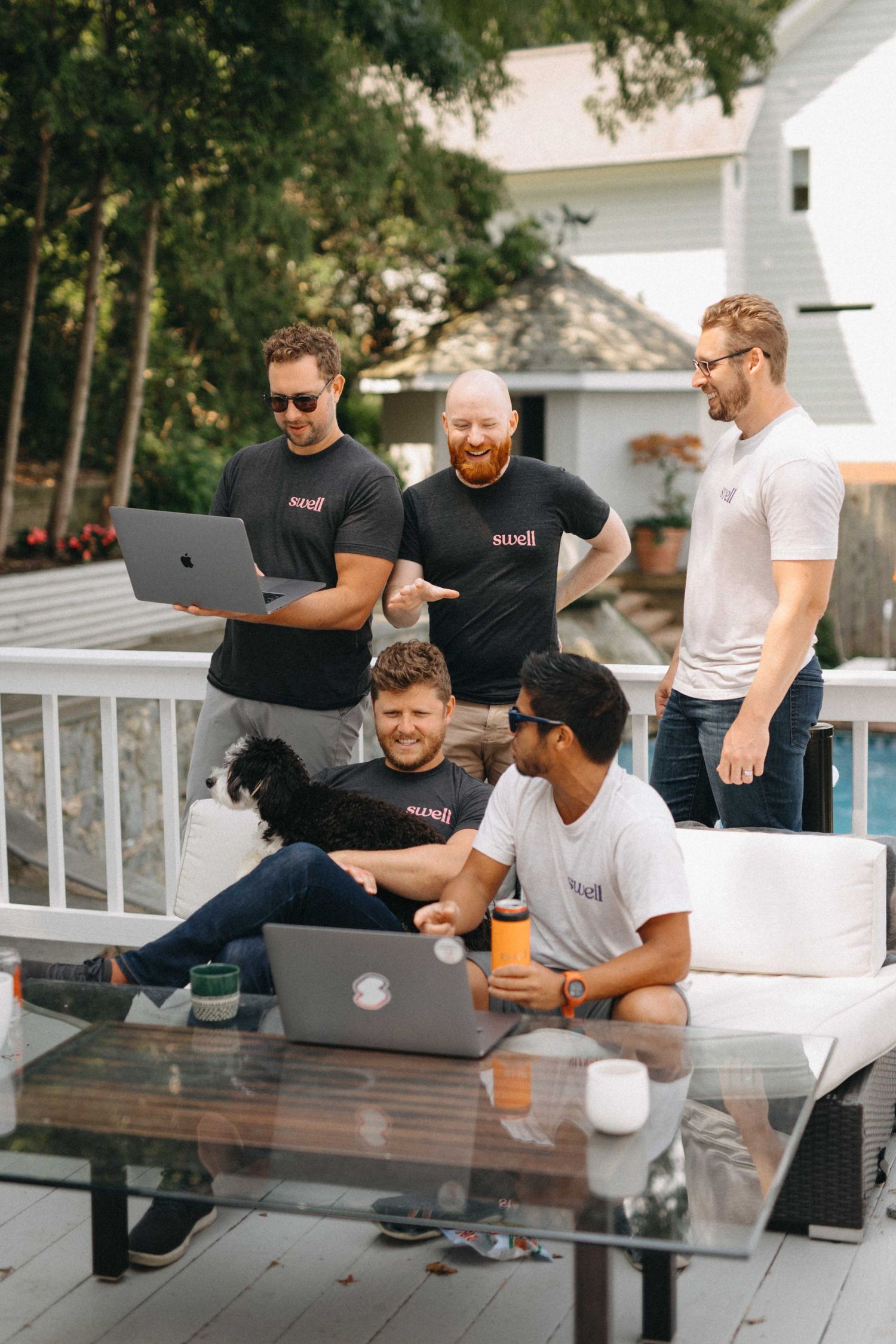Group of five men gathered around laptops on outdoor patio deck reviewing work together.