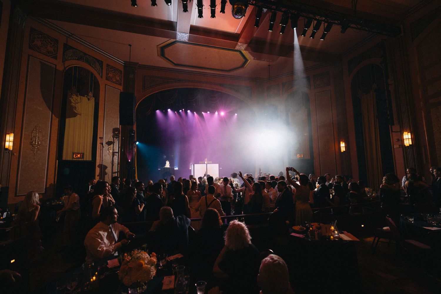 Wedding guests dance and celebrate on the floor of a grand ballroom with stage lighting.