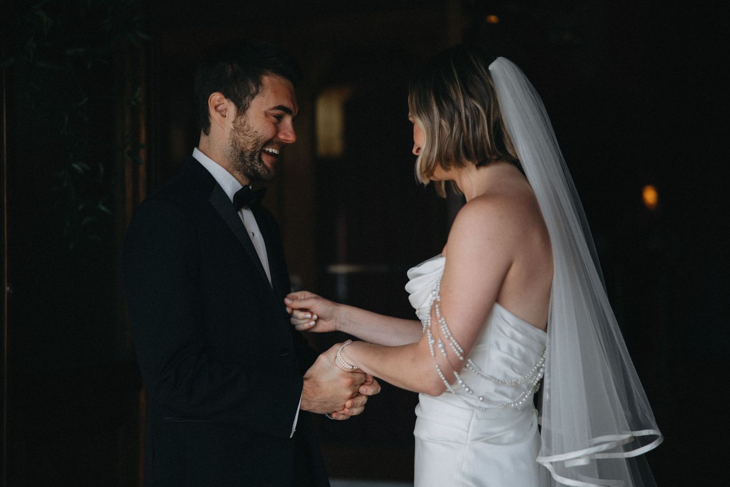 Bride and groom sharing joyful first look moment holding hands in dimly lit venue.