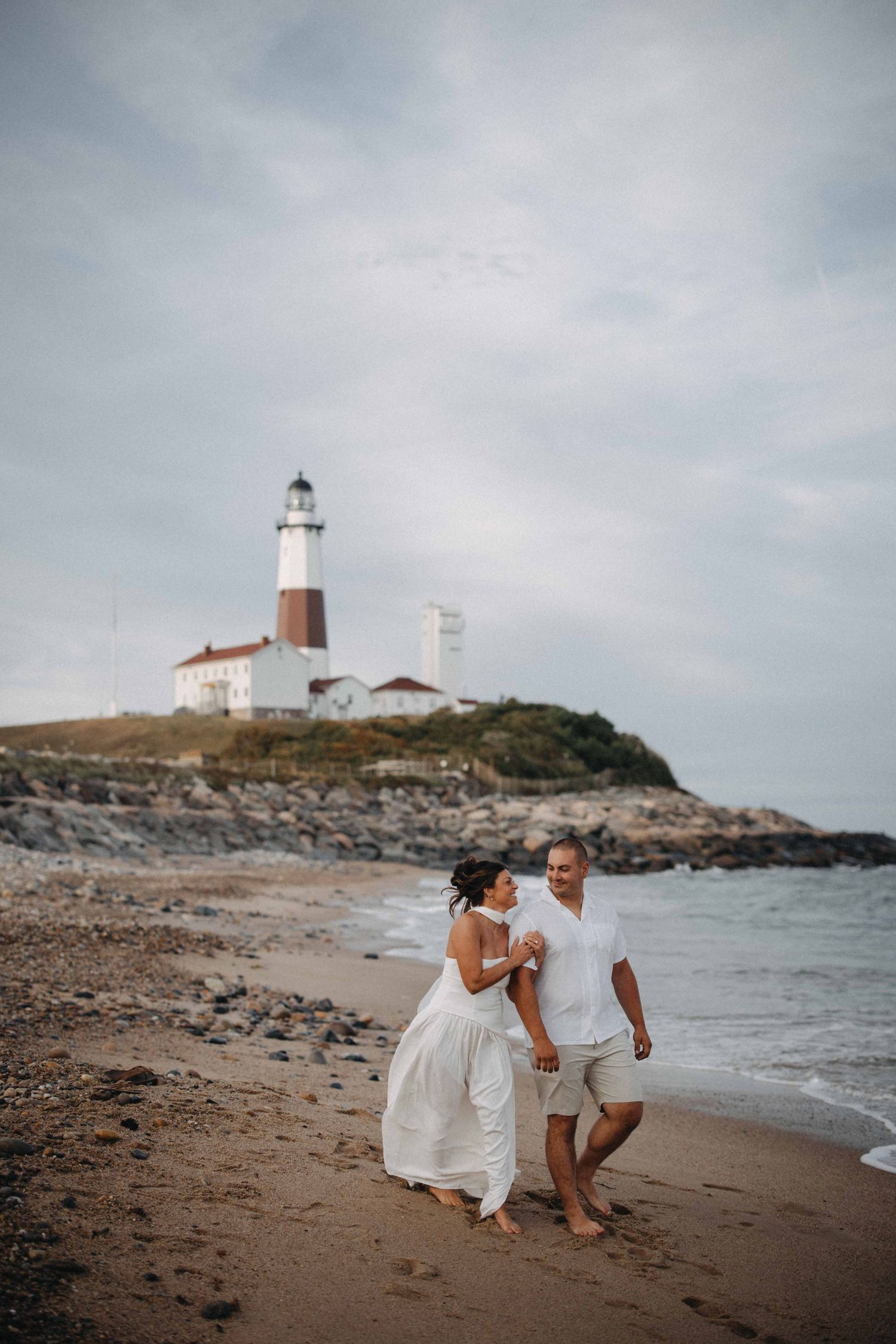 Couple in white attire walking hand in hand on a beach with a lighthouse behind.