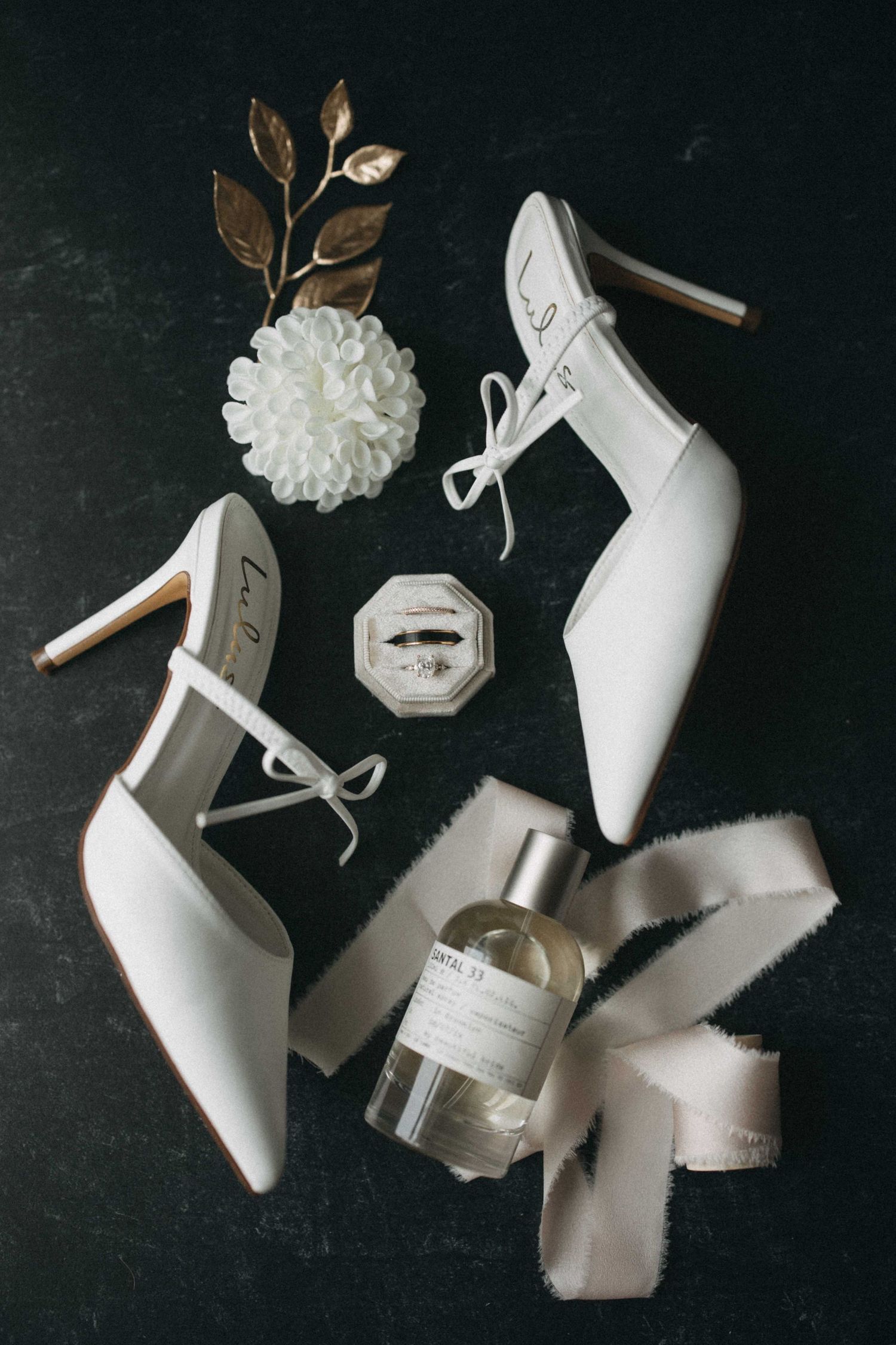 Flat lay of white bridal heels, ring box, perfume bottle, and floral accessories on dark surface.