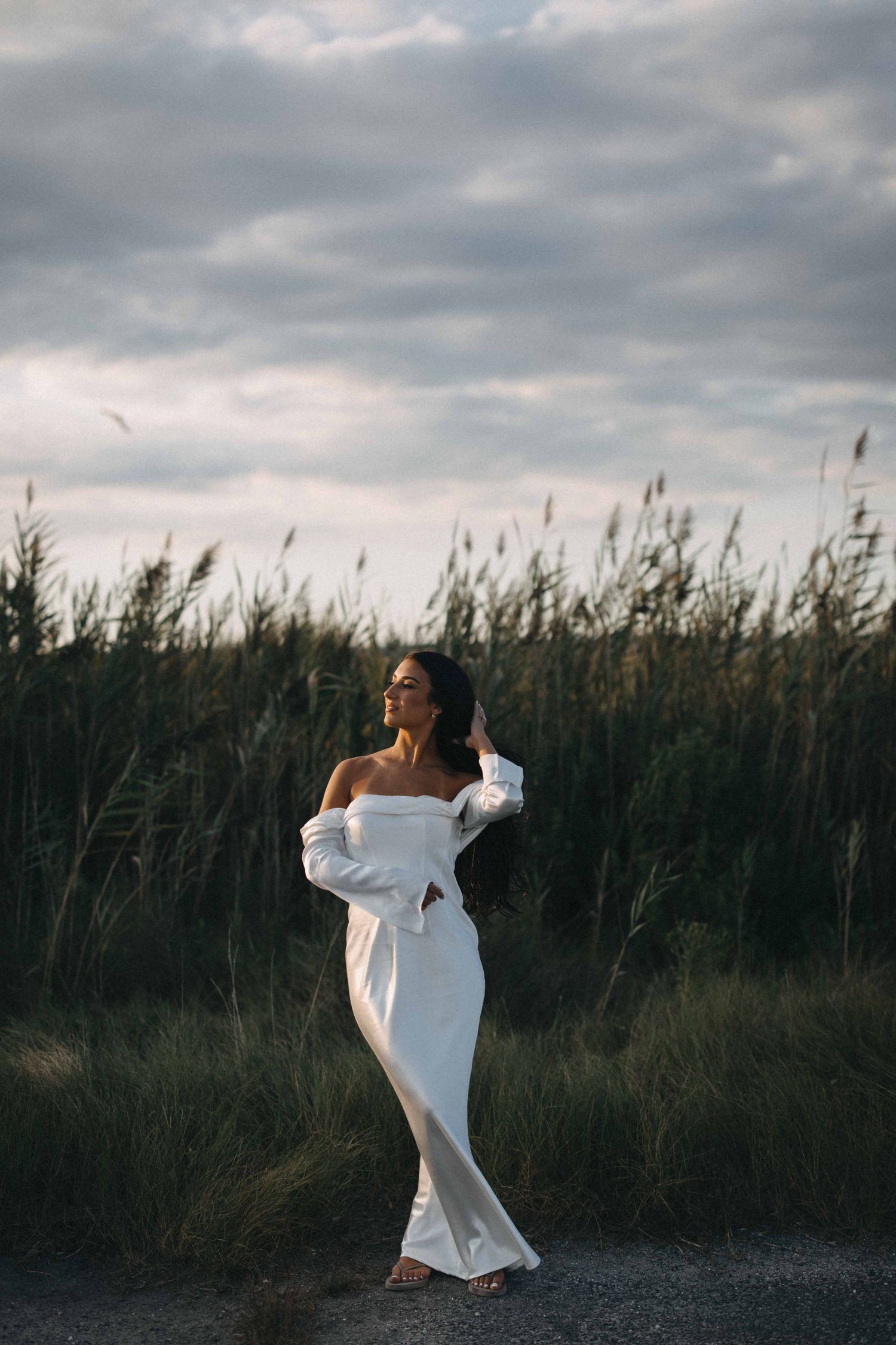 Bride in an off-shoulder white gown posing gracefully in a field of tall grass under cloudy skies.