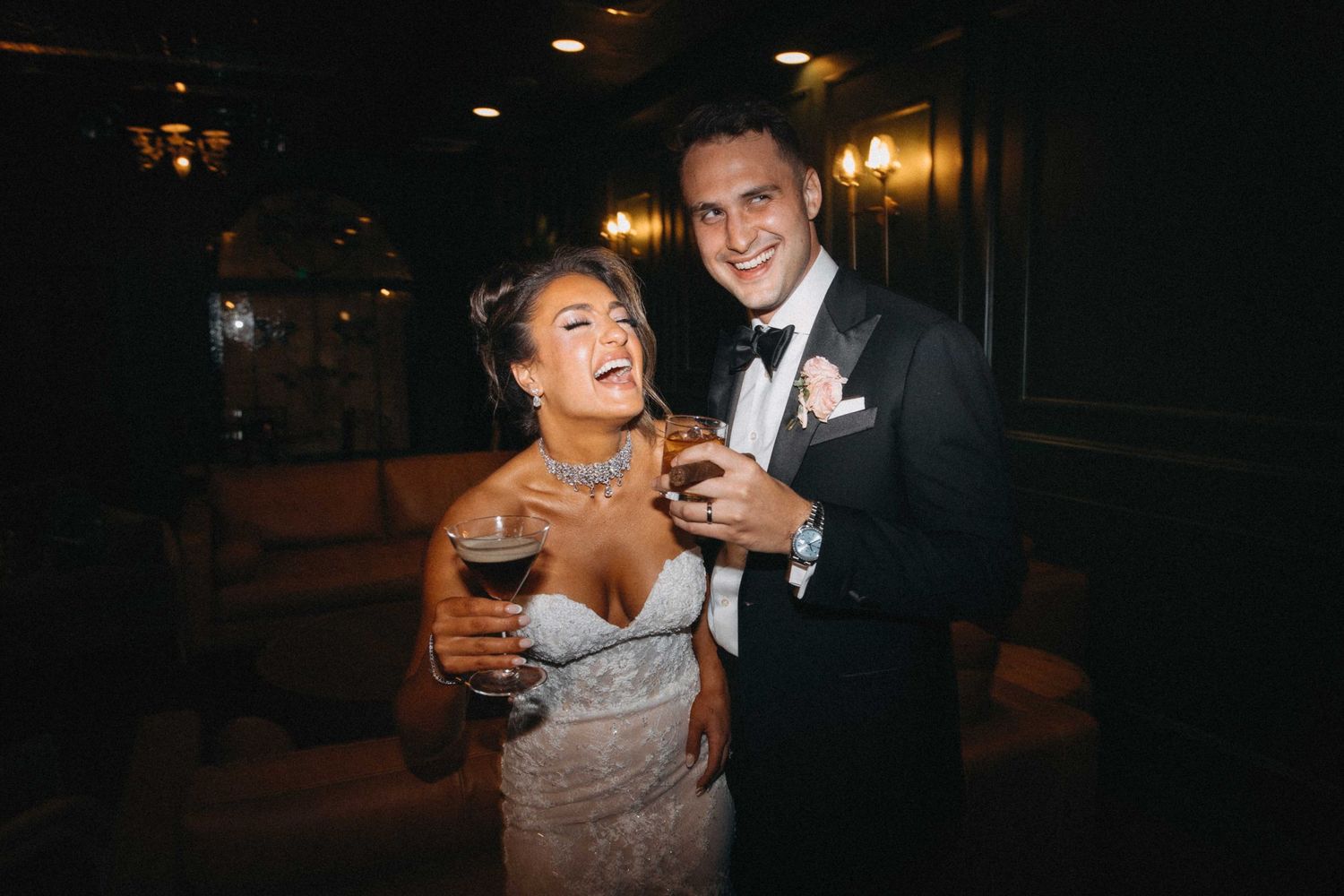 Happy bride and groom laughing together while holding drinks at their wedding reception in a dimly lit venue.