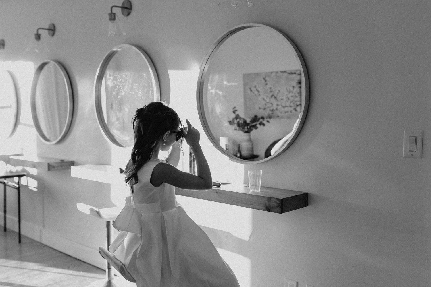 Black and white photo of bride adjusting hair at vanity with round mirrors.