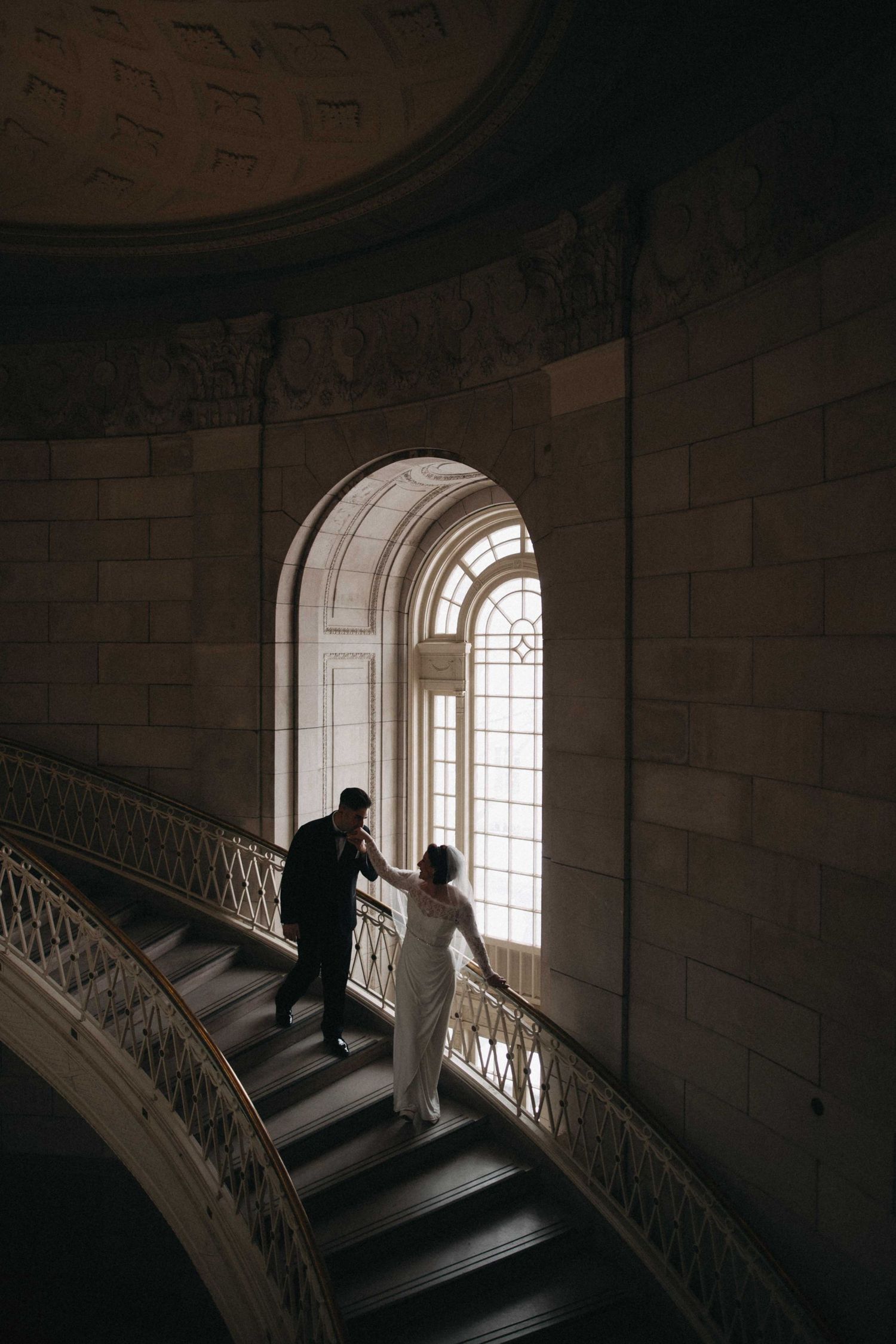 Bride and groom silhouetted on a grand curved staircase before a large arched window in a historic building.