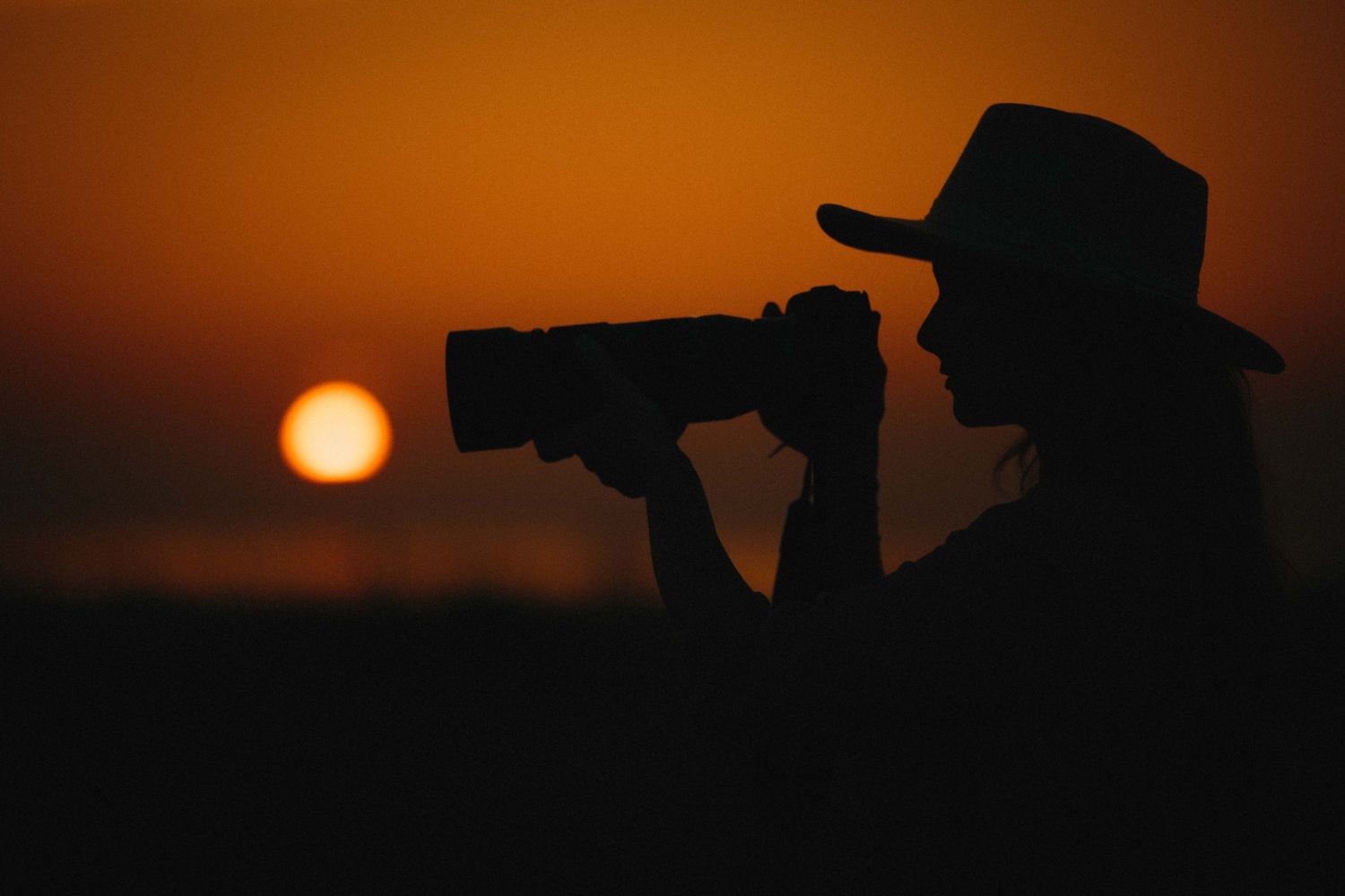 Silhouette of a woman in a hat photographing a vibrant orange sunset with a telephoto camera lens.