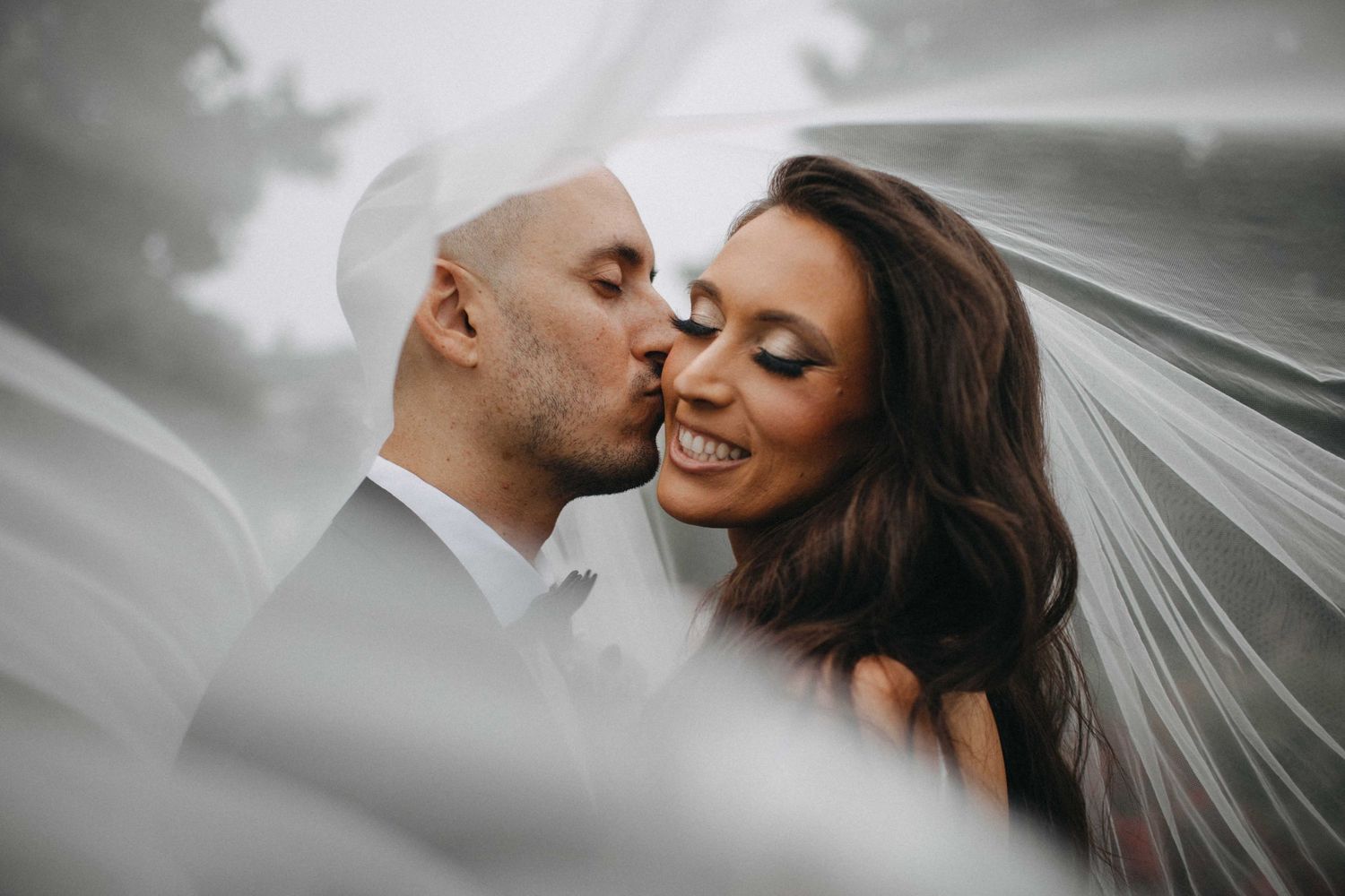 Romantic bride and groom portrait with flowing veil, groom kissing bride's cheek as she smiles joyfully.