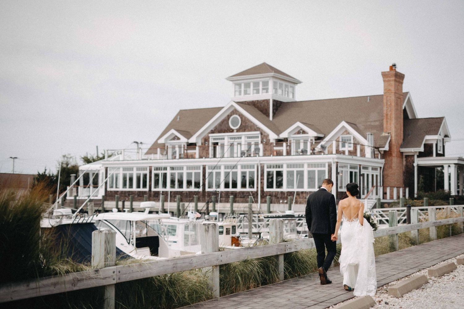 Bride and groom walking hand in hand along a wooden dock toward a large waterfront wedding venue.
