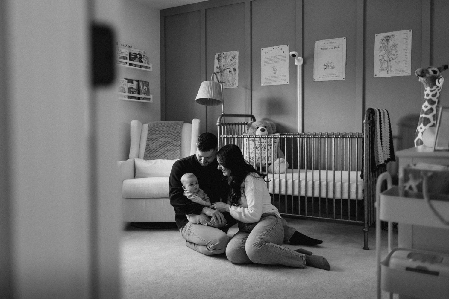 Black and white photo of parents sitting on nursery floor cradling newborn baby near crib.