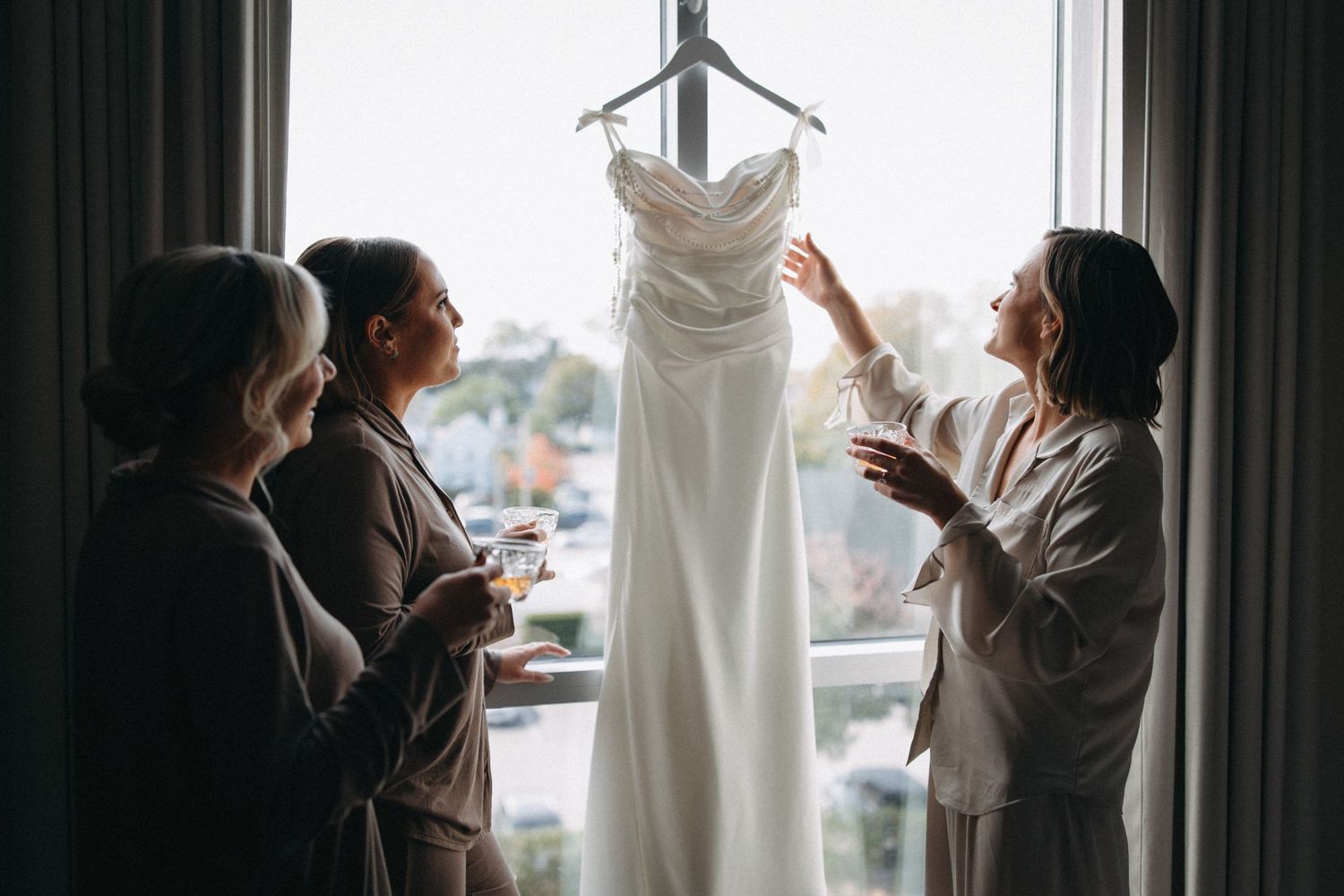 Bridesmaids in robes admiring white wedding gown hanging by window with champagne glasses.