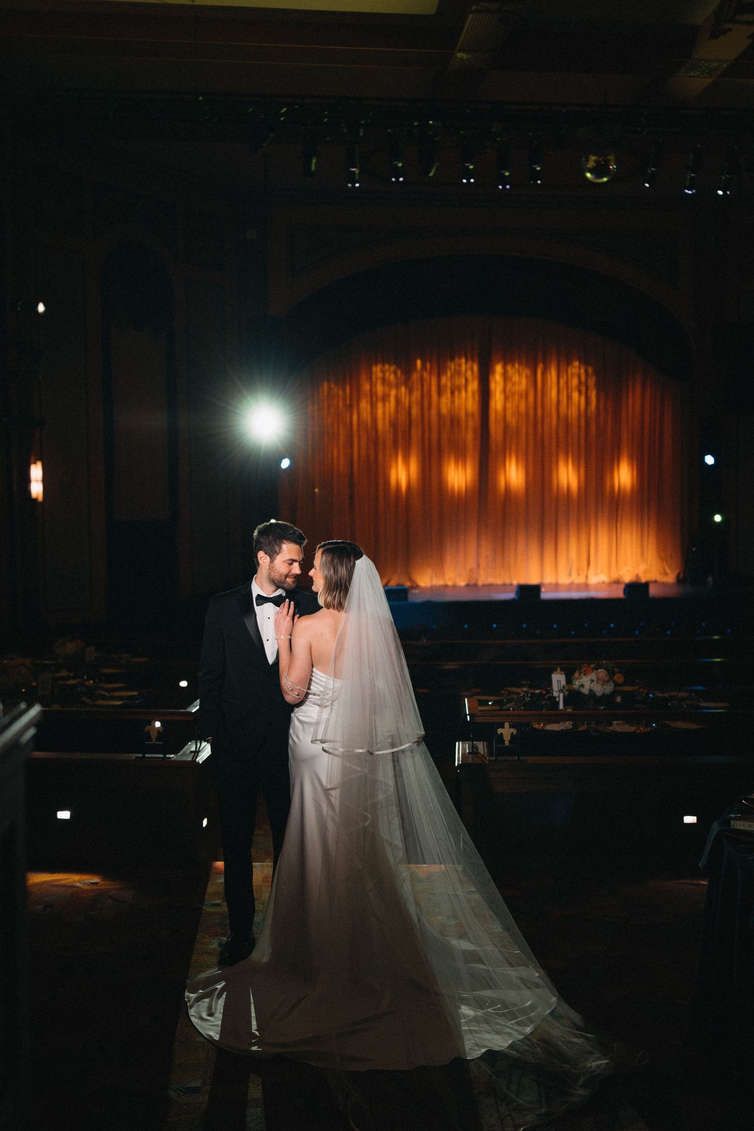 Bride and groom share romantic moment in dimly lit historic theater venue with golden stage curtain.