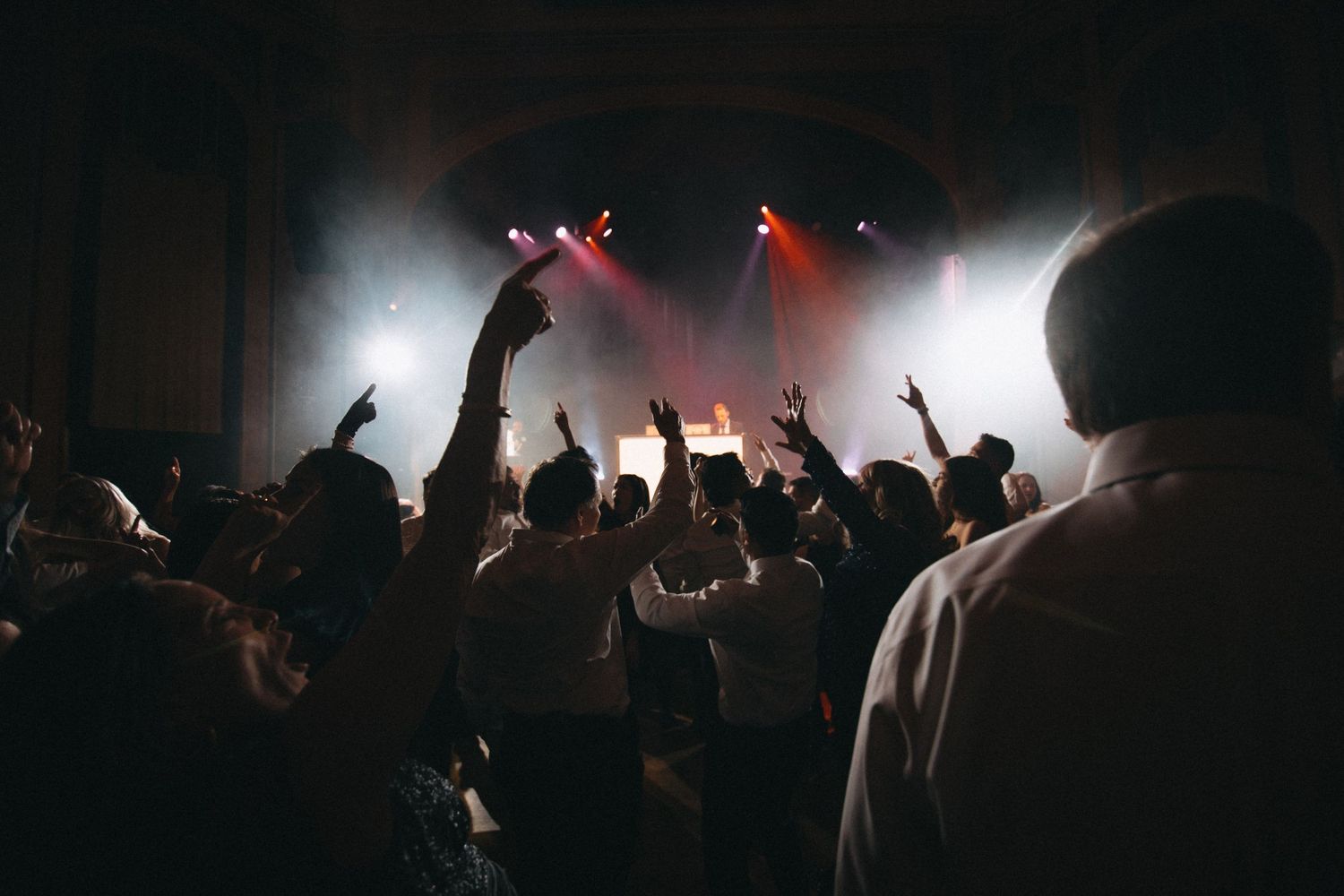 Energetic wedding reception dance floor crowd with raised hands under colorful stage lighting.