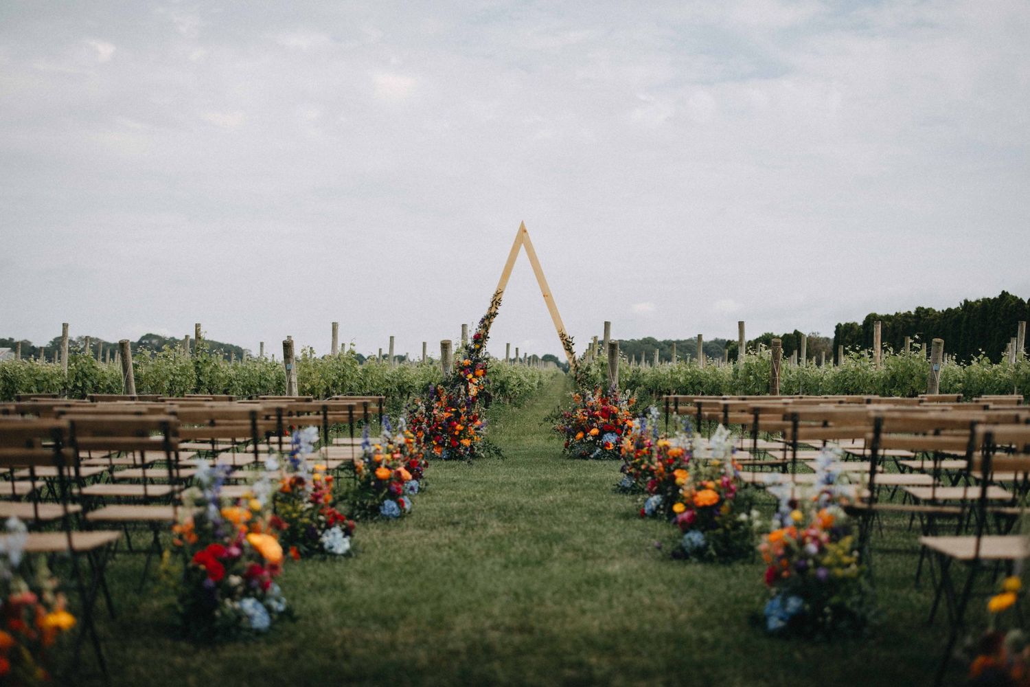 Outdoor vineyard wedding ceremony aisle with colorful floral arrangements and triangular arch.