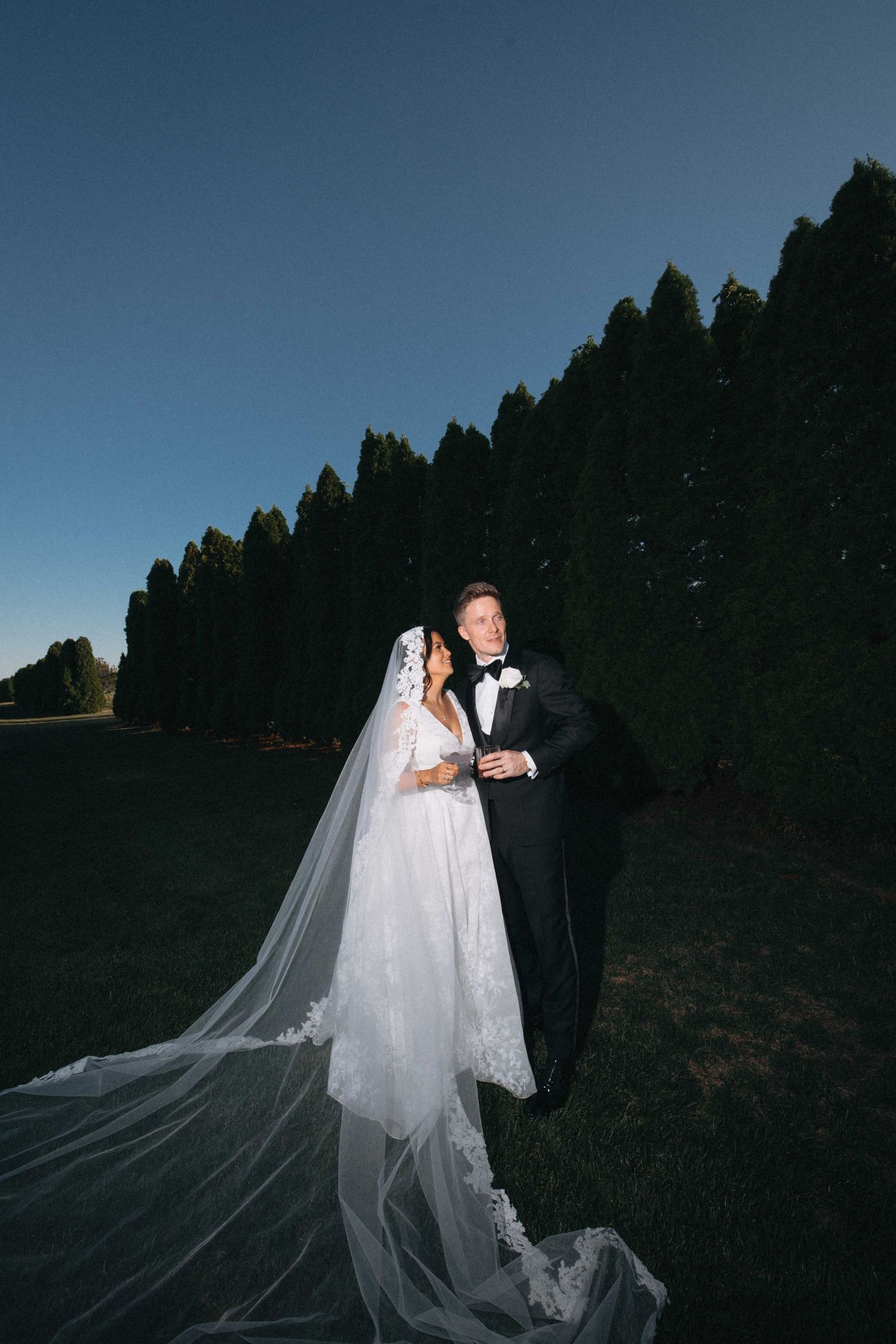 Bride in lace gown with long veil and groom in tuxedo at dusk.