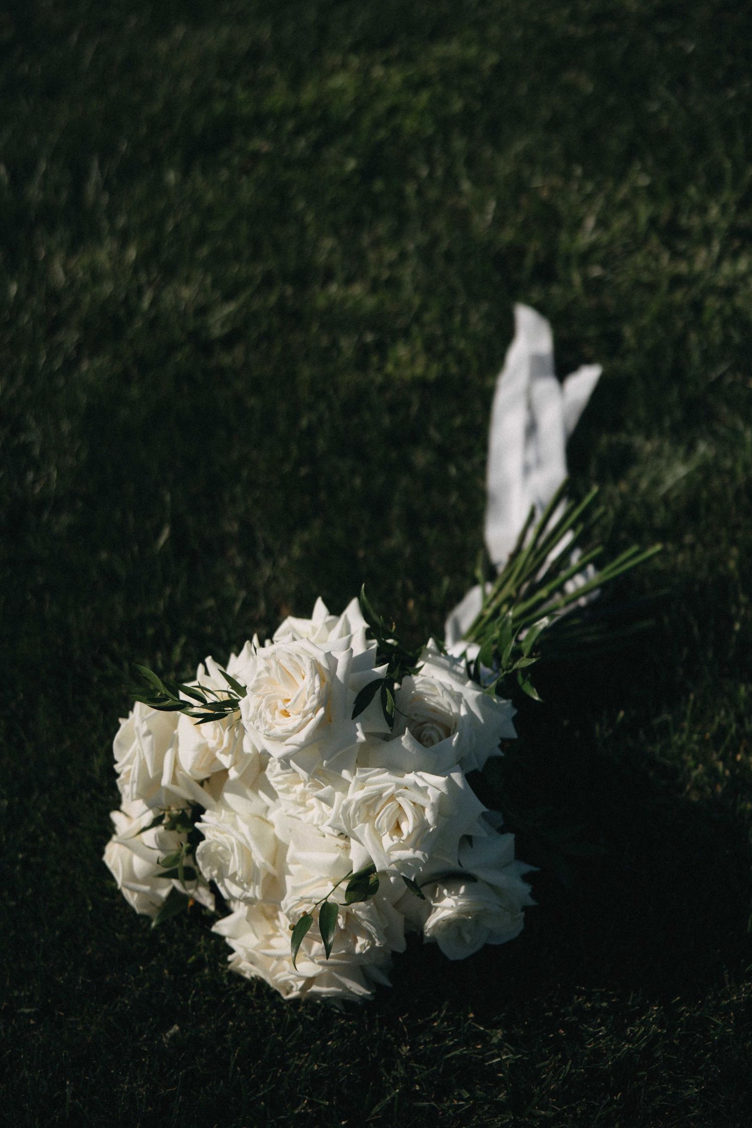 Elegant white rose bridal bouquet with ribbon resting on green grass.