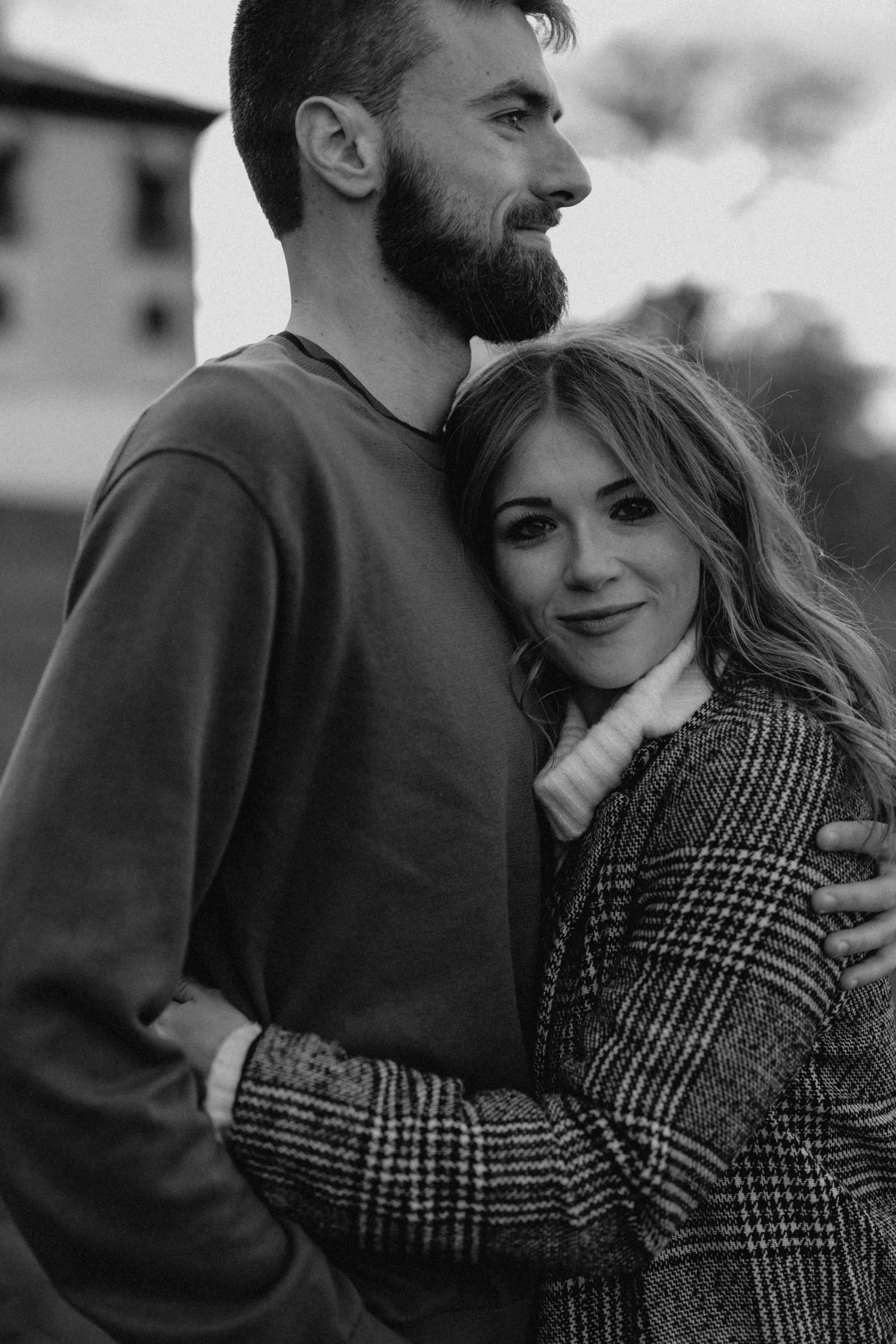 Black and white portrait of a bearded man embracing a smiling woman outdoors.