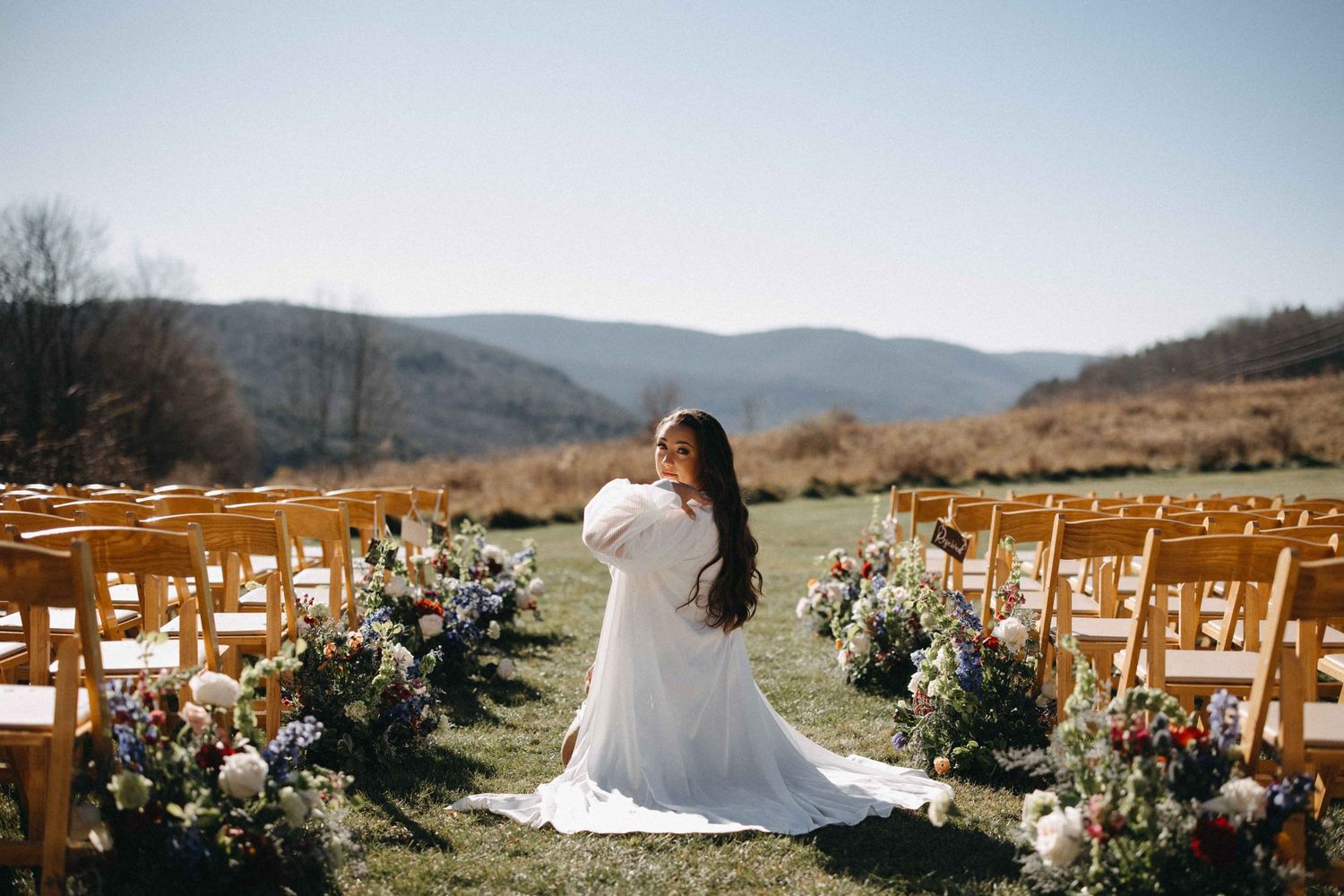 Bride in white gown kneeling among wildflower arrangements at an outdoor mountain wedding ceremony venue.