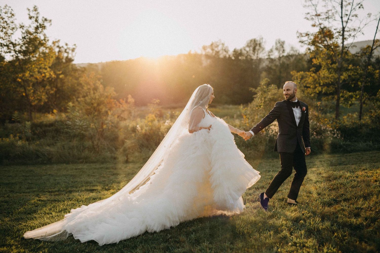 Bride and groom walking through sunlit meadow at golden hour, bride in voluminous white gown.