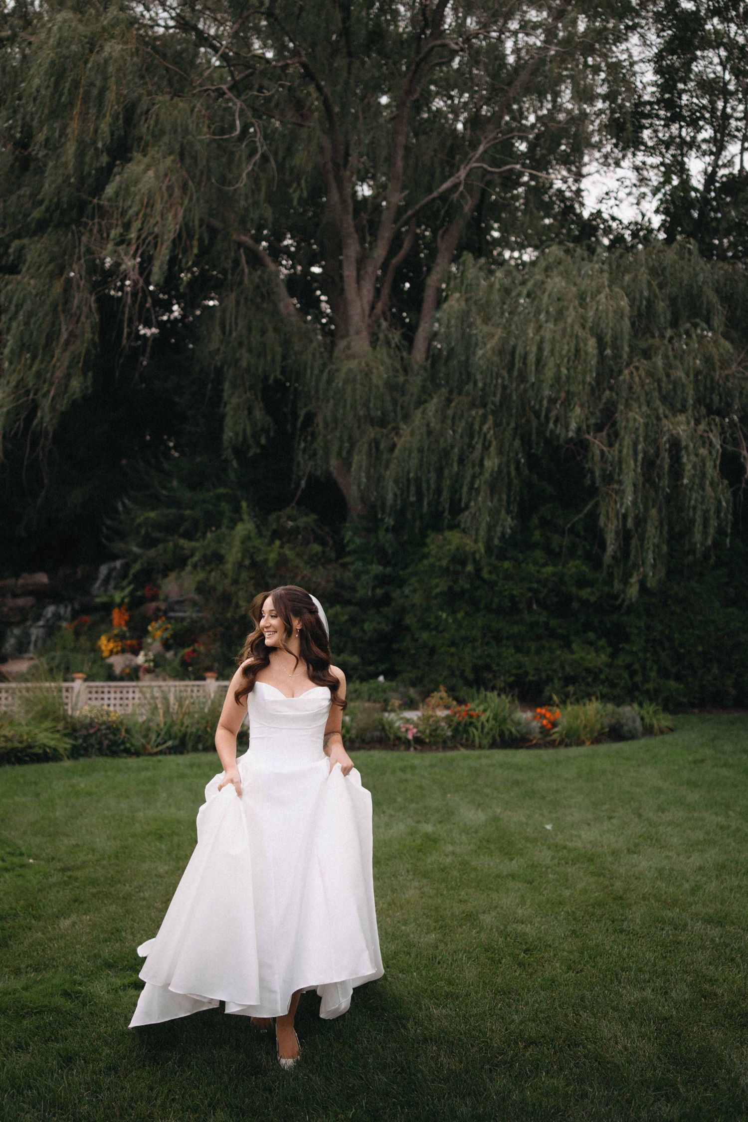 Bride in strapless white gown walks through lush garden with weeping willow trees at dusk.