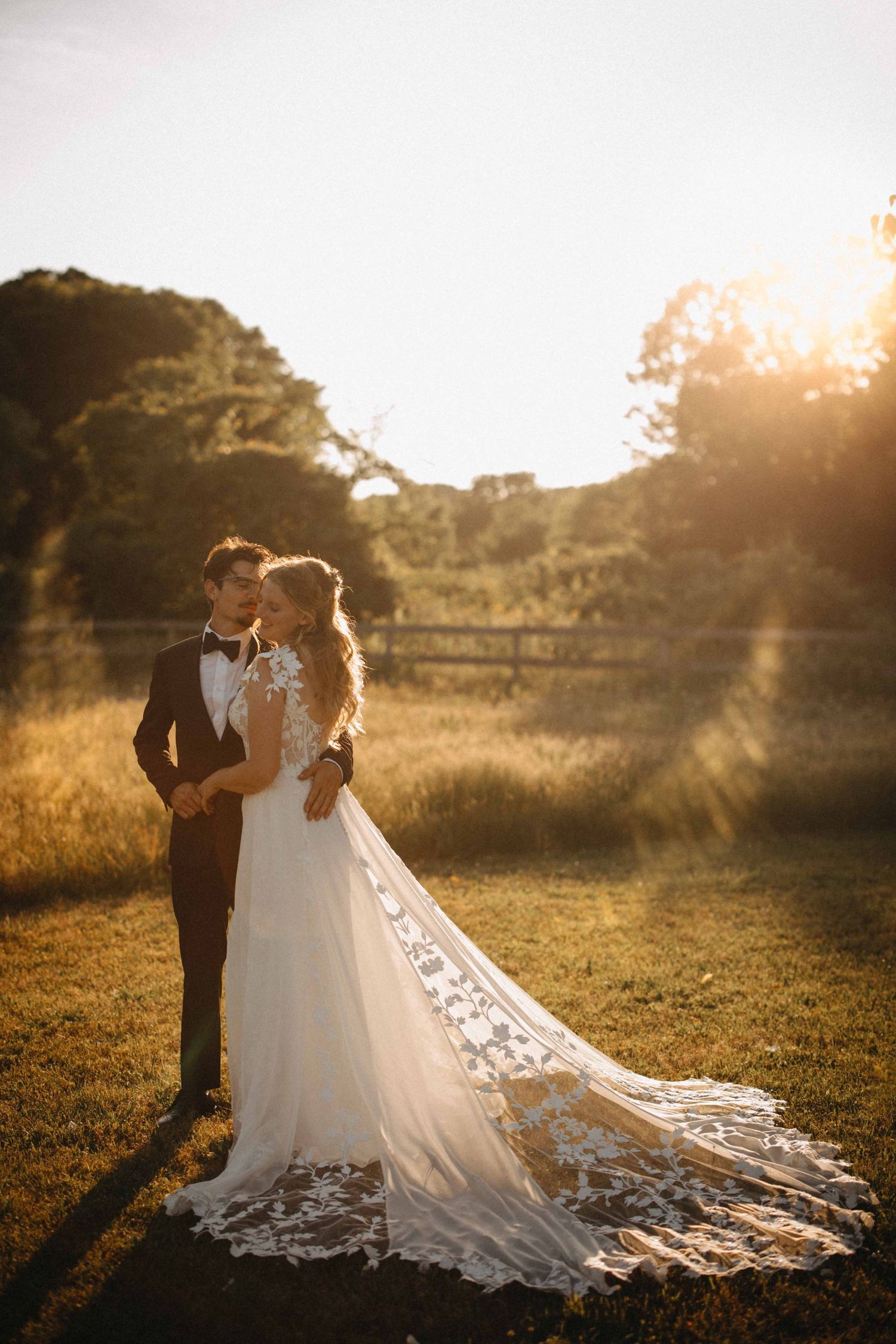 Bride and groom share a romantic golden hour moment in a sunlit field, her lace cathedral train spread beautifully on the grass.