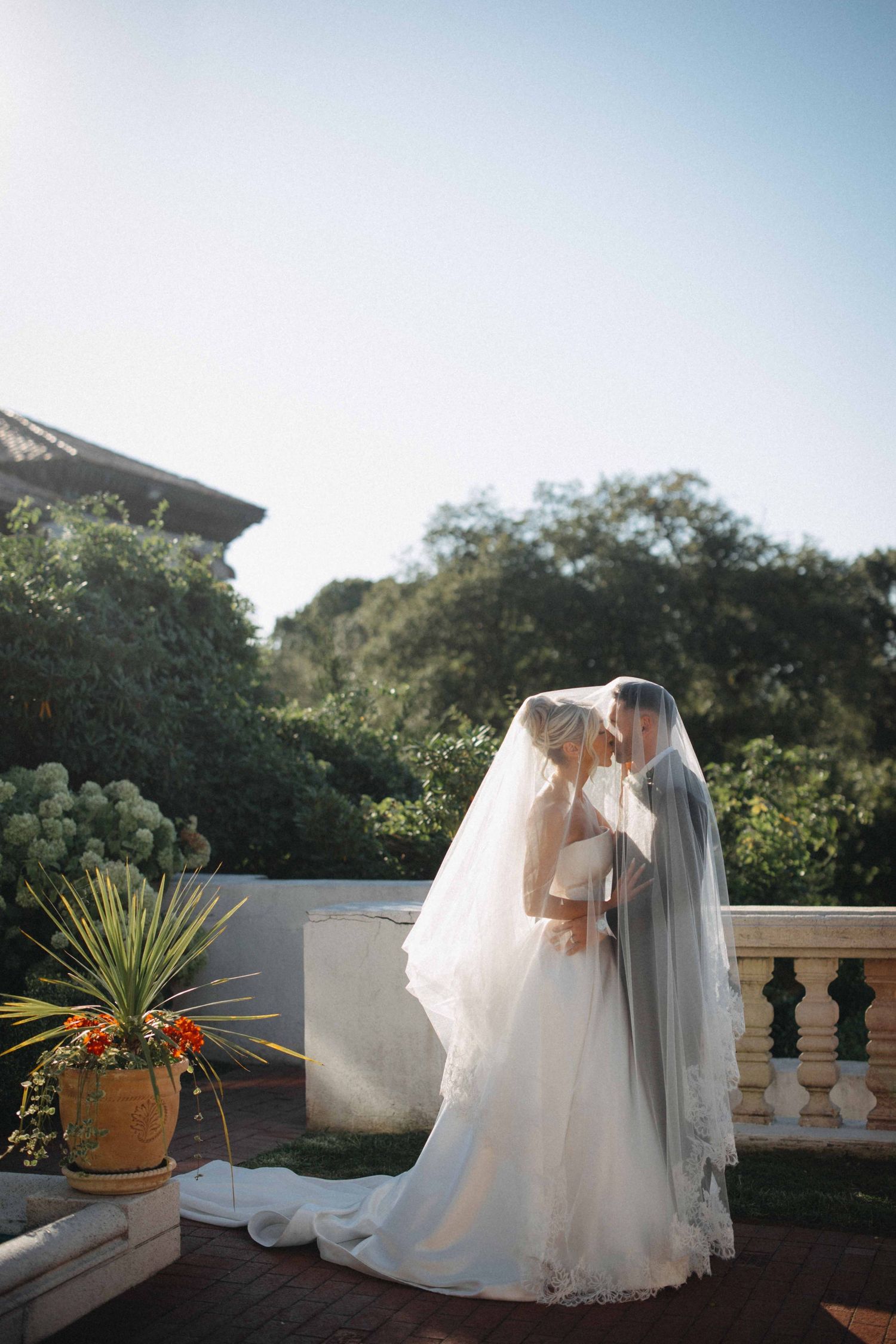 Bride and groom sharing a romantic kiss under a flowing veil on an elegant outdoor terrace surrounded by lush greenery.