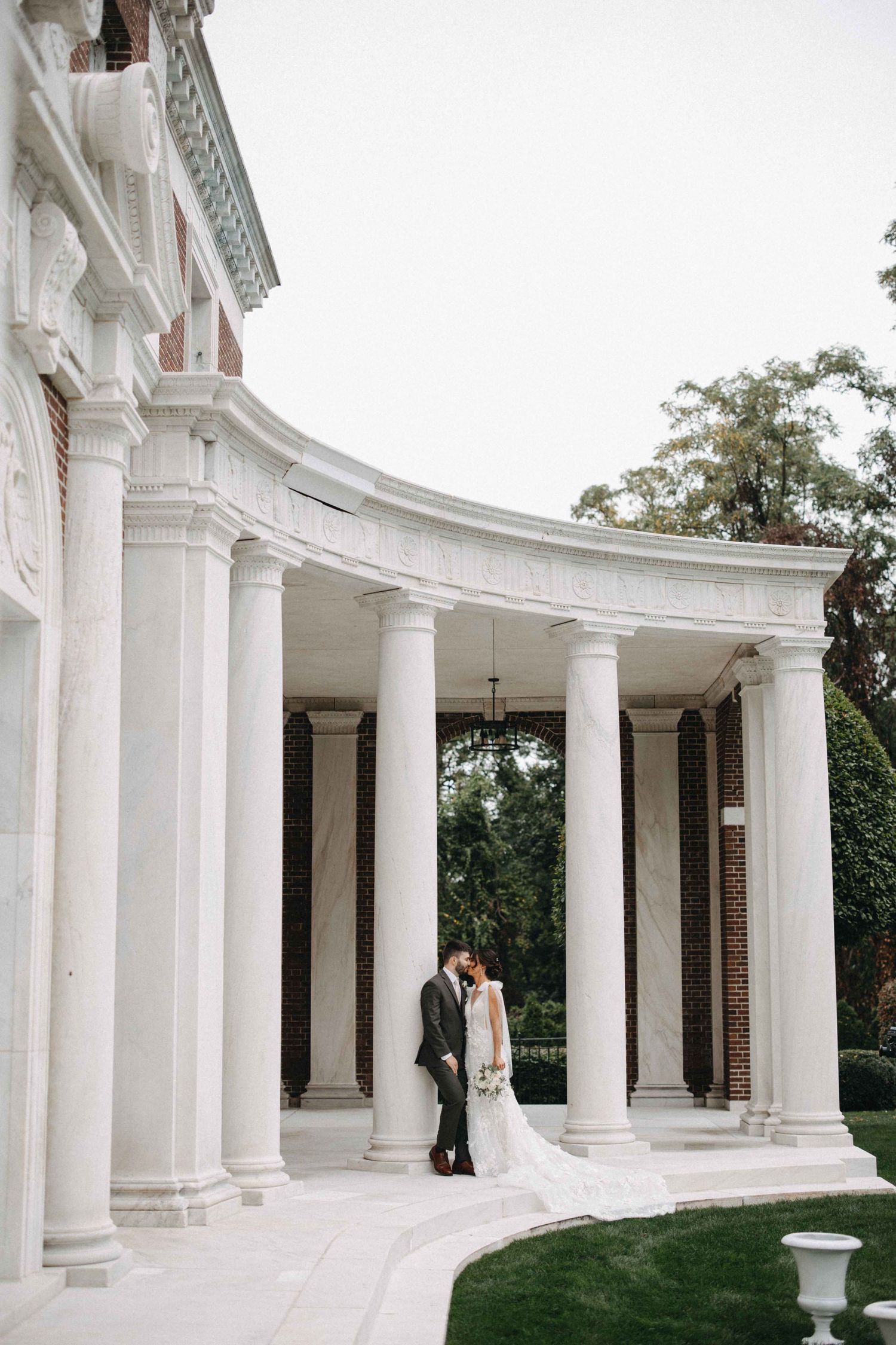 Bride and groom sharing a romantic kiss beneath grand white columns of a classic wedding venue.