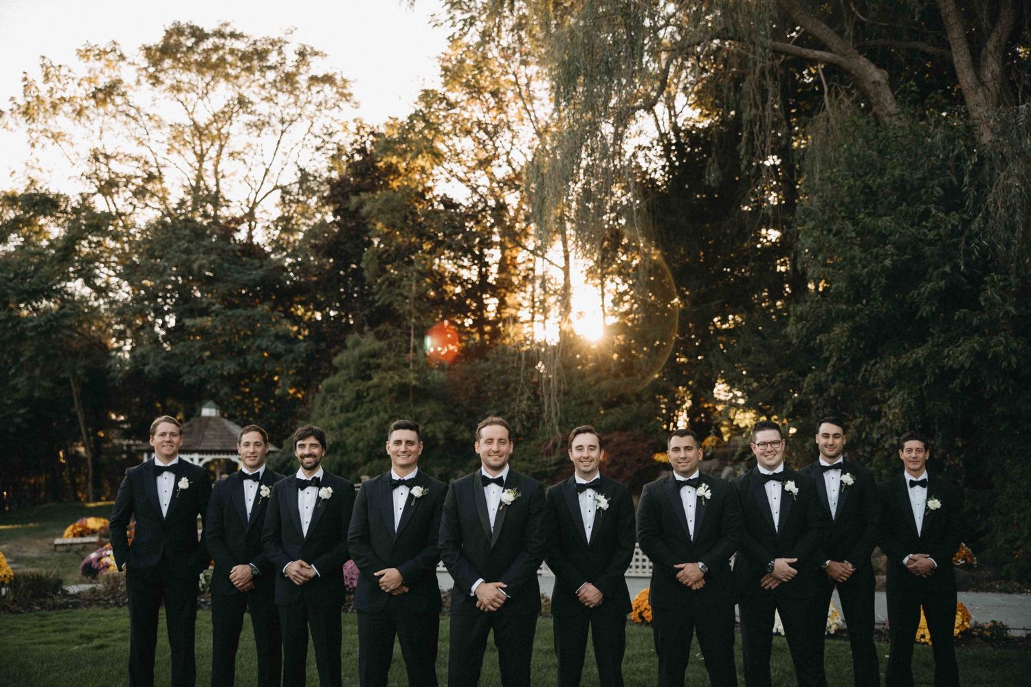 Groomsmen lined up in black tuxedos with bow ties at golden hour outdoor wedding.