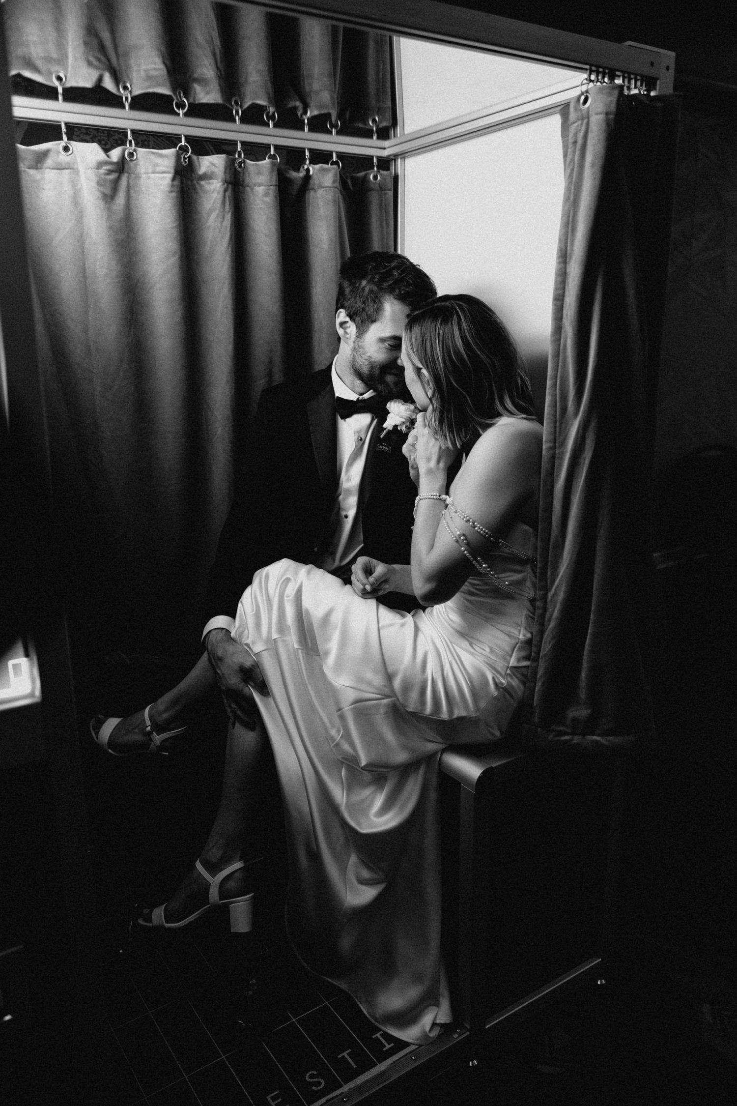 Bride and groom share an intimate moment together inside a vintage photo booth in black and white.